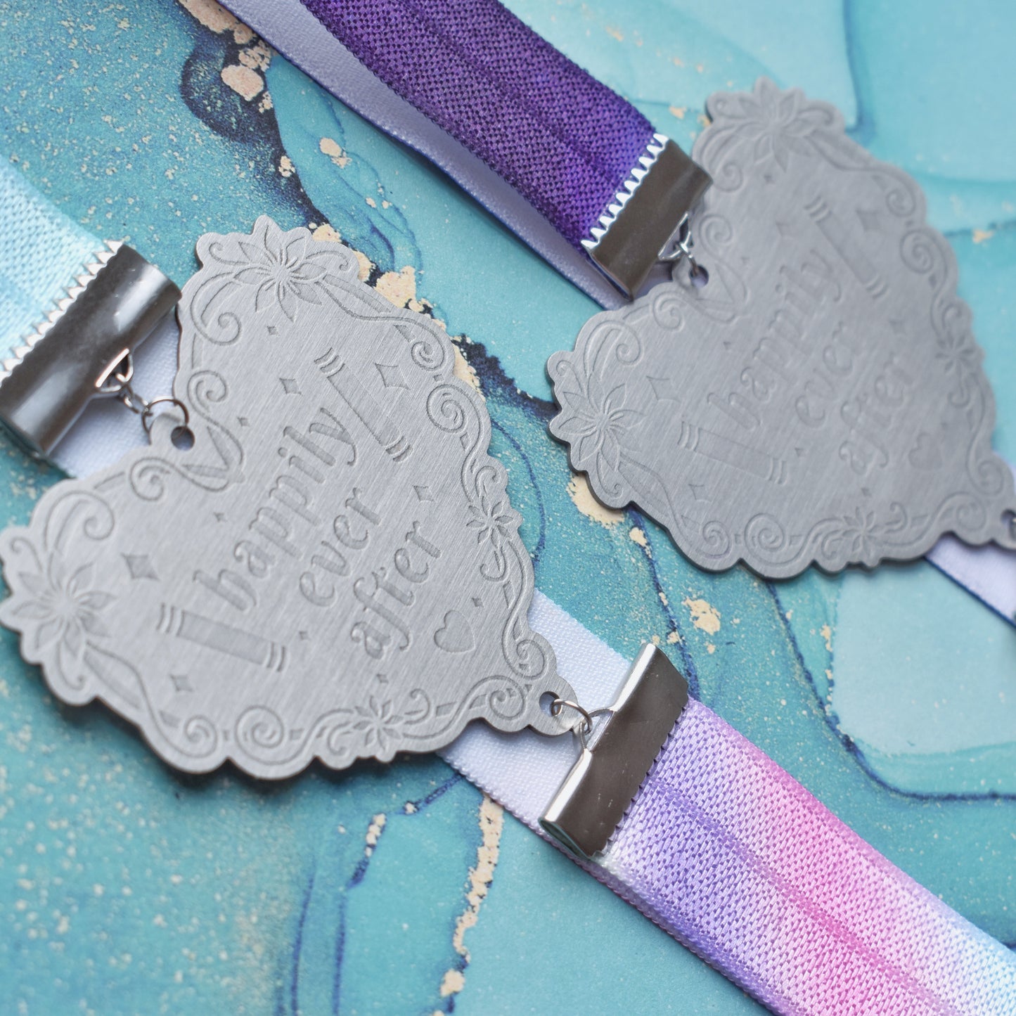 2 book bands at diagonal in the centre of image. Bottom bookband is pastel rainbow elastic with stainless steel engraved heart and book charm that reads happily ever after. Top bookband is purple galaxy elastic with stainless steel engraved heart and book charm that reads happily ever after. On a pale blue/teal watercolour background.