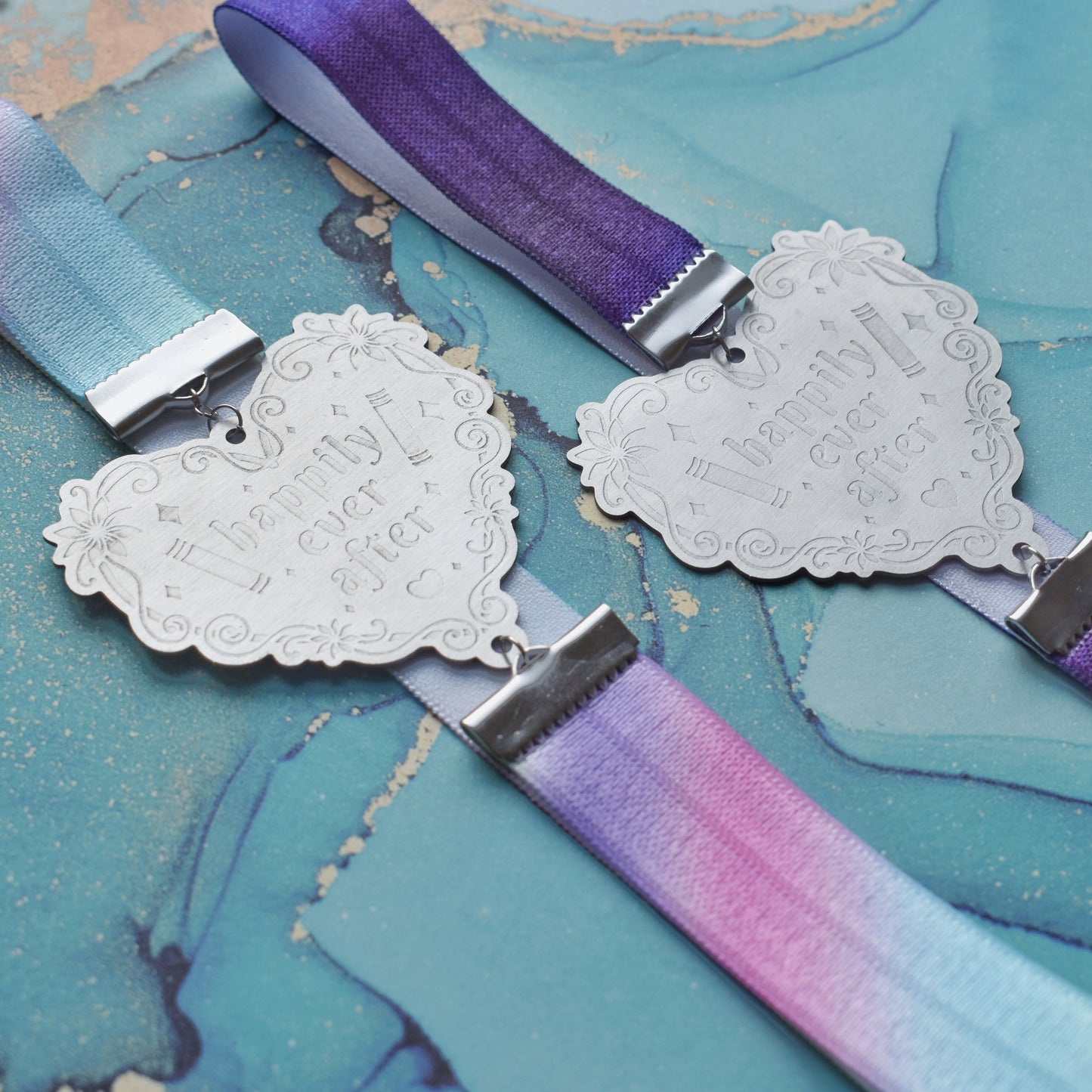 2 book bands at diagonal in the centre of image. Bottom bookband is pastel rainbow elastic with stainless steel engraved heart and book charm that reads happily ever after. Top bookband is purple galaxy elastic with stainless steel engraved heart and book charm that reads happily ever after. On a pale blue/teal watercolour background.