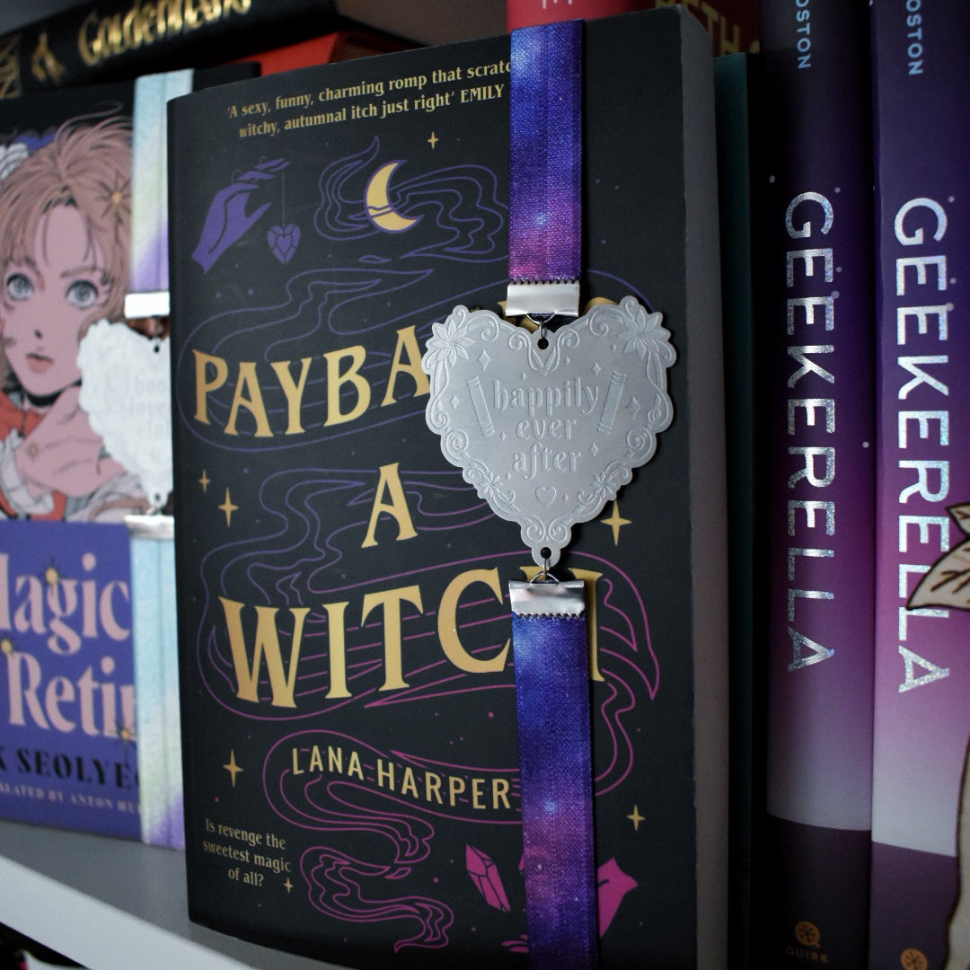 Book titled Payback's A Witch is shown with a book band wrapped around top to bottom on right hand side. The bookband is purple galaxy elastic with stainless steel engraved heart and book charm that reads happily ever after. Bookshelf and other books can be seen in background and forground of image.