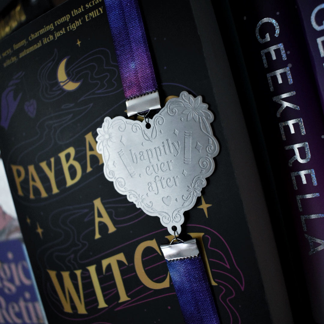 Close up of book titled Payback's A Witch is shown with a book band wrapped around top to bottom on right hand side. The bookband is purple galaxy elastic with stainless steel engraved heart and book charm that reads happily ever after. Bookshelf and other books can be seen in background and forground of image.
