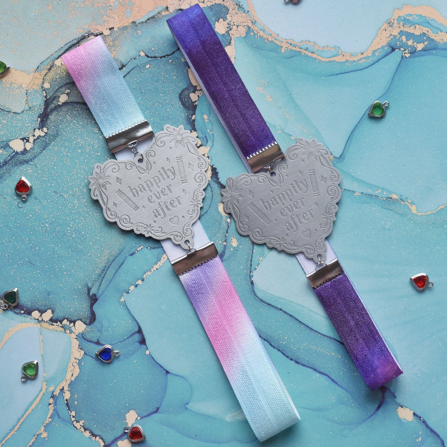 2 book bands at slight diagonal in the centre of image. Left bookband is pastel rainbow elastic with stainless steel engraved heart and book charm that reads happily ever after. Right bookband is purple galaxy elastic with stainless steel engraved heart and book charm that reads happily ever after. On a pale blue/teal watercolour bacground with silver and colour gem hearts scattered around.