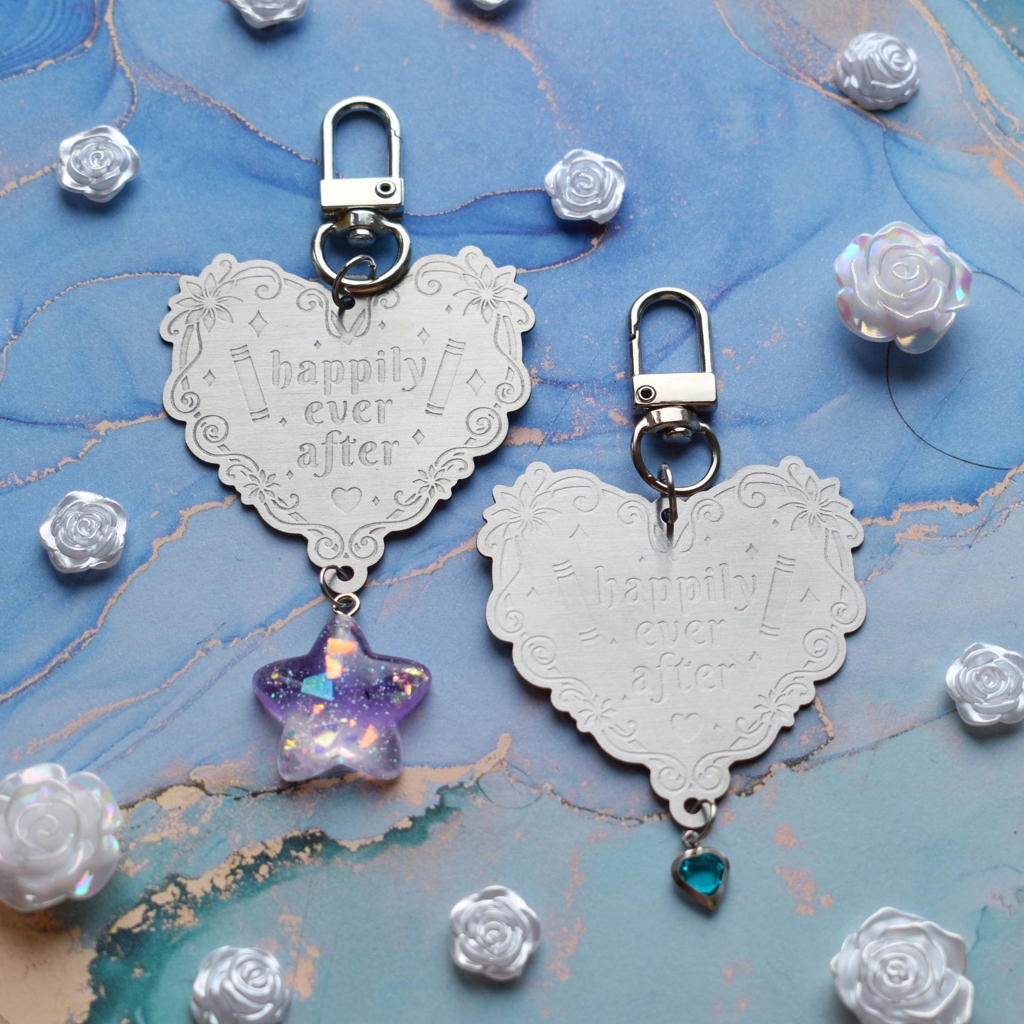 2 keyrings with stainless steel engraved heart and book charm that reads happily ever after in the centre of image. Left keyring has a lilac glittery cabachon star attached to charm. Right keyring has a small silver and gem pale blue heart attached to charm. On a pale blue/teal watercolour background with white rose cabachons scattered around.
