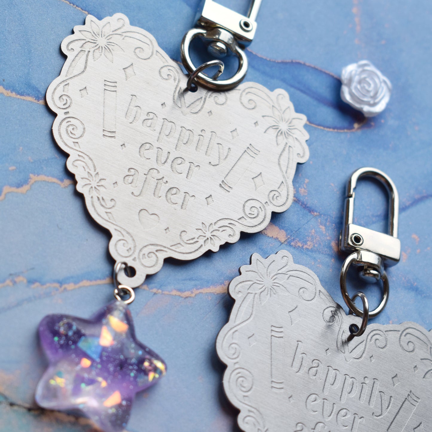 Close up of 2 keyrings with stainless steel engraved heart and book charm that reads happily ever after at diagonal in the centre of image. Left keyring has a lilac glittery cabachon star attached to charm. Right keyring has a small silver and gem pale blue heart attached to charm. On a pale blue/teal watercolour background with white rose cabachons scattered around.