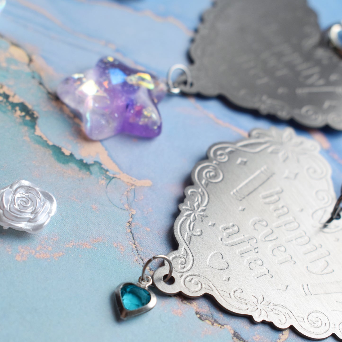 Close up of 2 keyrings with stainless steel engraved heart and book charm that reads happily ever after at diagonal in the centre of image. Left keyring has a lilac glittery cabachon star attached to charm. Right keyring has a small silver and gem pale blue heart attached to charm. On a pale blue/teal watercolour background with white rose cabachons scattered around.
