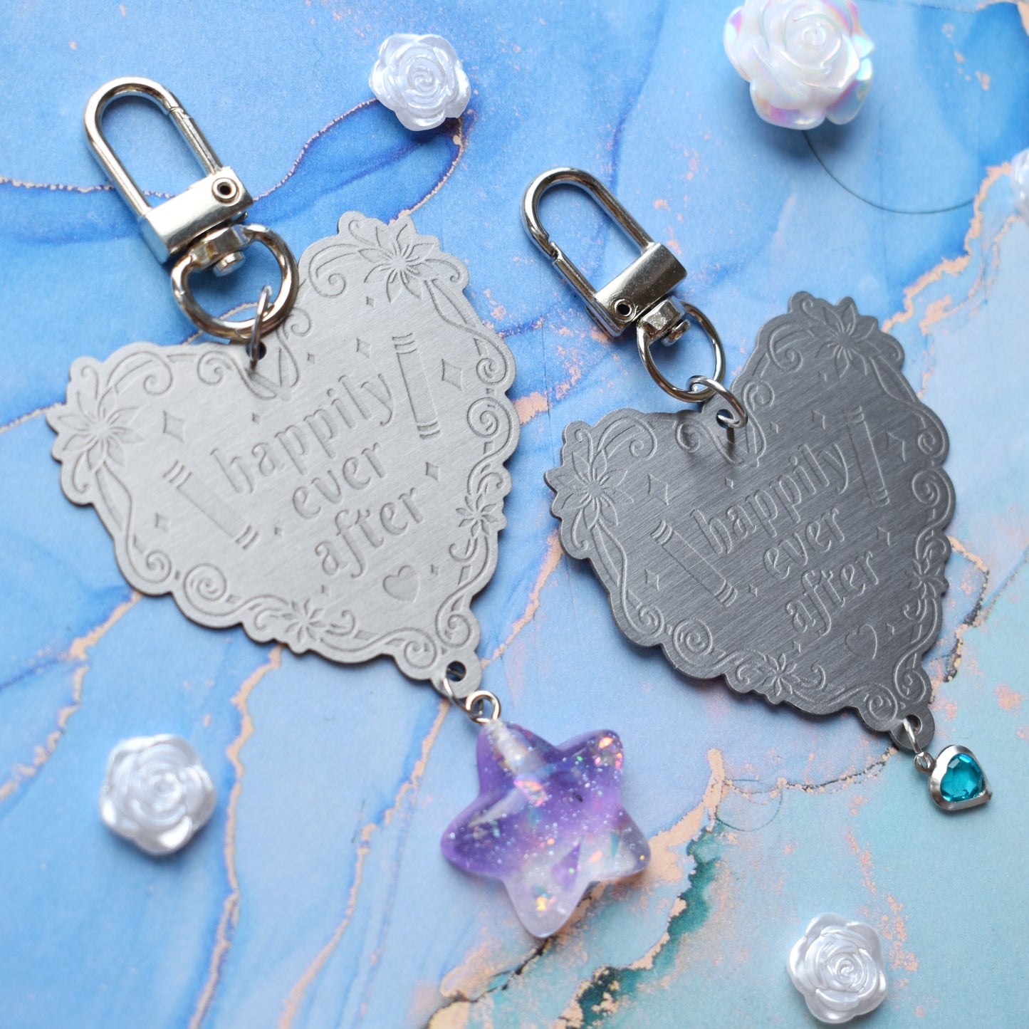 2 keyrings with stainless steel engraved heart and book charm that reads happily ever after at diagonal in the centre of image. Left keyring has a lilac glittery cabachon star attached to charm. Right keyring has a small silver and gem pale blue heart attached to charm. On a pale blue/teal watercolour background with white rose cabachons scattered around.