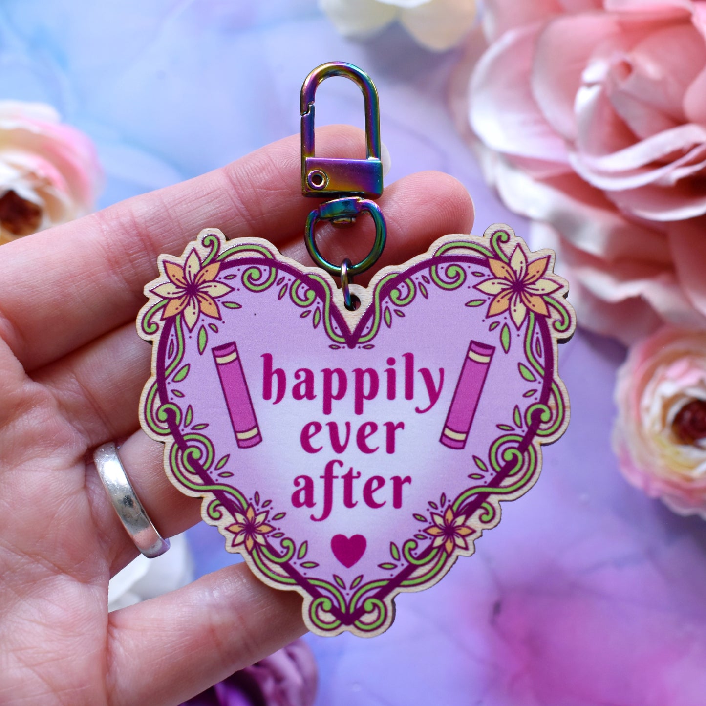 Pink heart-shaped bookish keyring version, design features text 'happily ever after' with a floral and swirly outline with book spines. Keyring held by a white hand above a watercolour style background in blue,pink and purples with flowers around the edge of the photo.