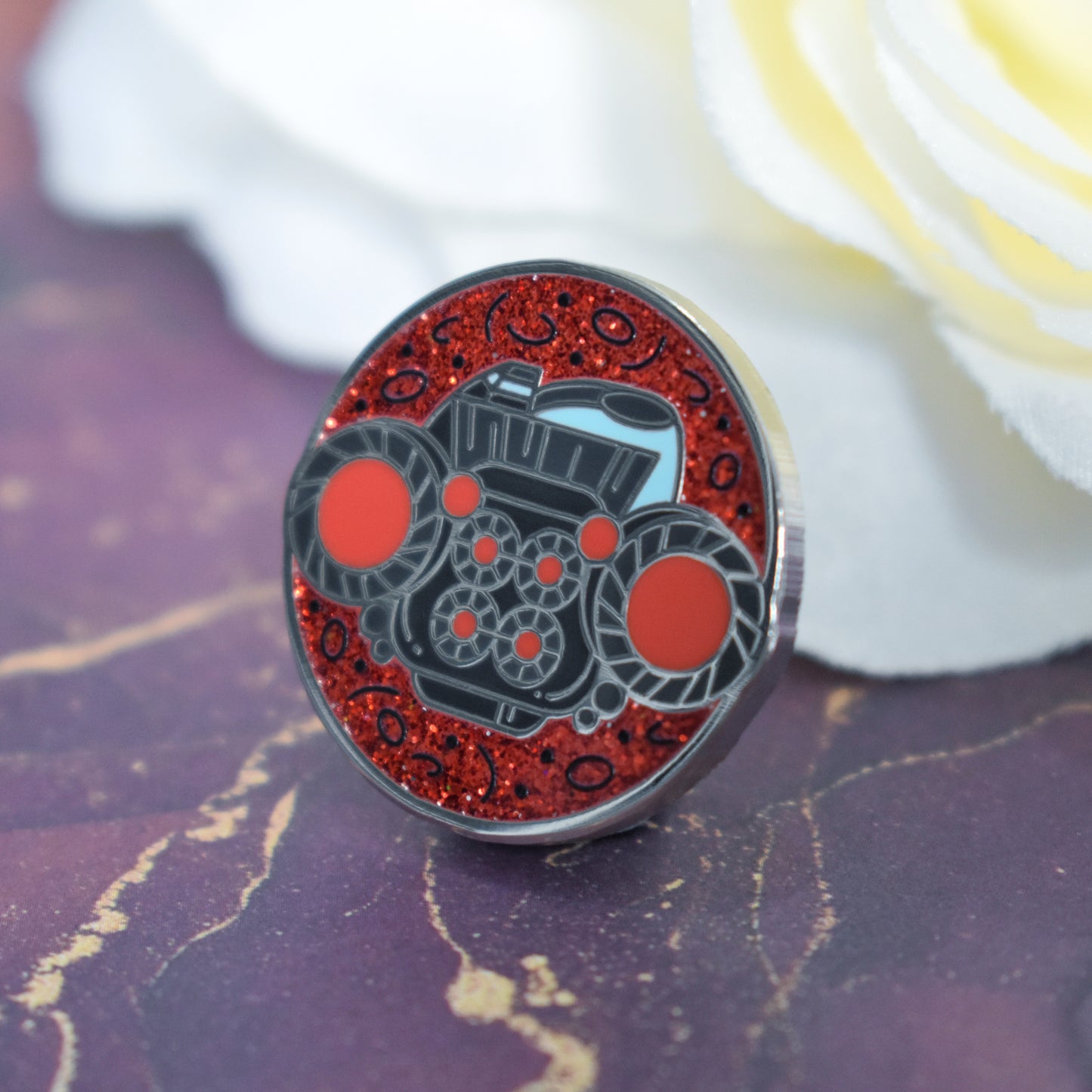 Silver metal enamel pin in centre of image. Enamel pin is round with a red glitter with tiny black detailed background visible on top and bottom. In the center of the design is the back end of a dark grey/blue blocky spaceship with red glowing engines. Background of photograph is dark pink/purple water inspired with a fake cream flower.