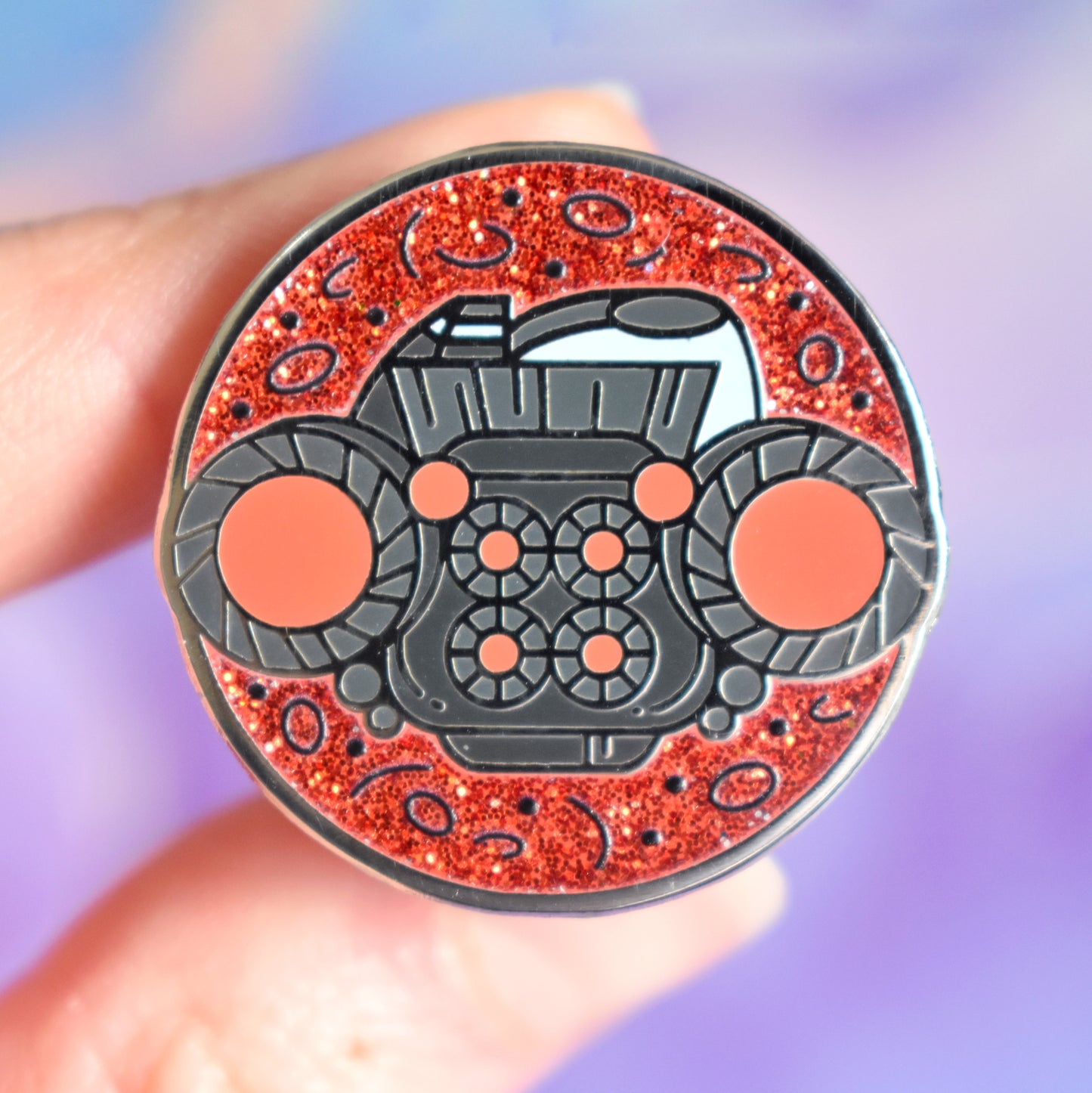 White hand holding silver metal enamel pin in centre of image. Enamel pin is round with a red glitter with tiny black detailed background visible on top and bottom. In the center of the design is the back end of a dark grey/blue blocky spaceship with red glowing engines. Background of photograph is dark pink/purple water inspired with a fake cream flower.