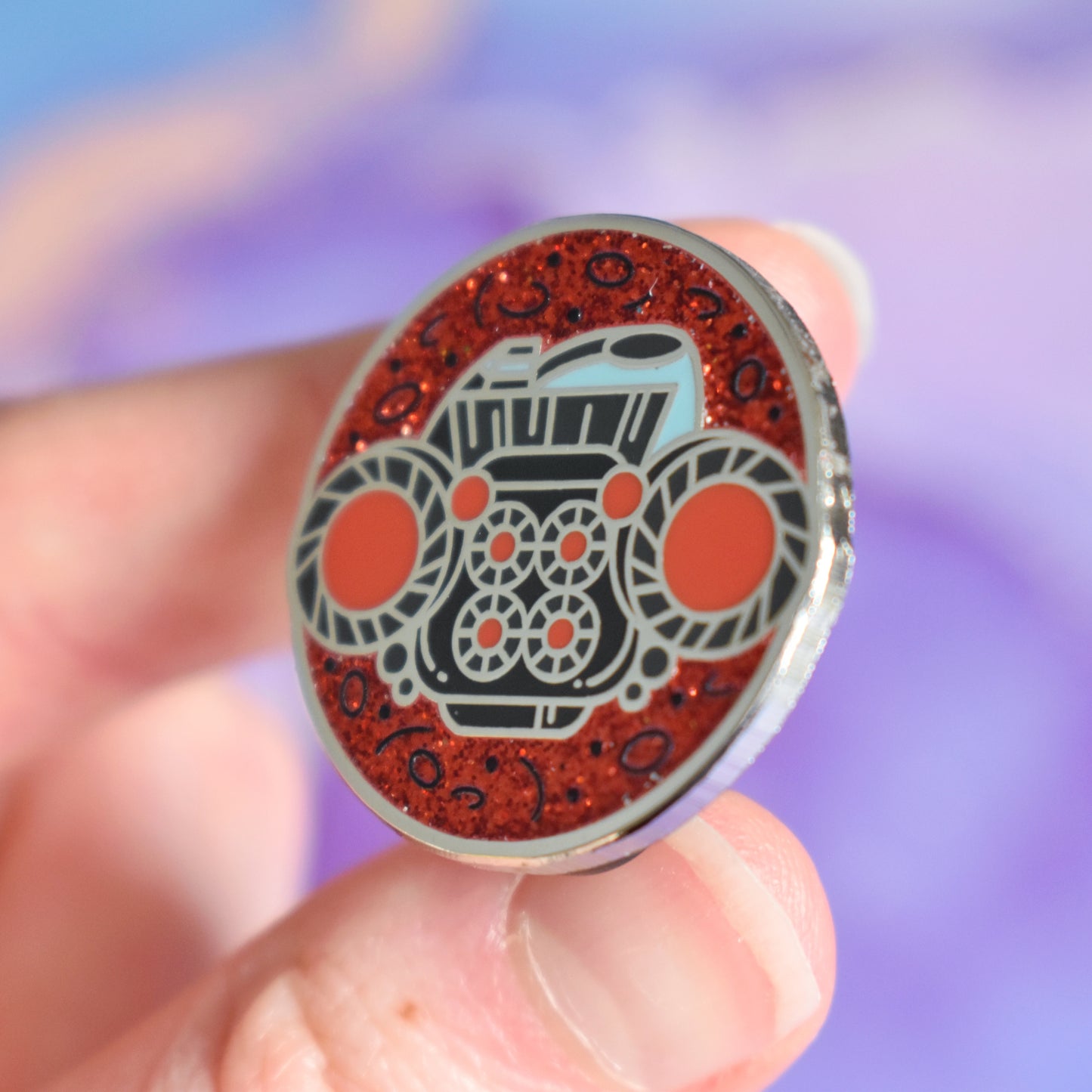 White hand holding silver metal enamel pin tilted to make the metal catch more light. Enamel pin is round with a red glitter with tiny black detailed background visible on top and bottom. In the center of the design is the back end of a dark grey/blue blocky spaceship with red glowing engines. Background of photograph is dark pink/purple water inspired with a fake cream flower.