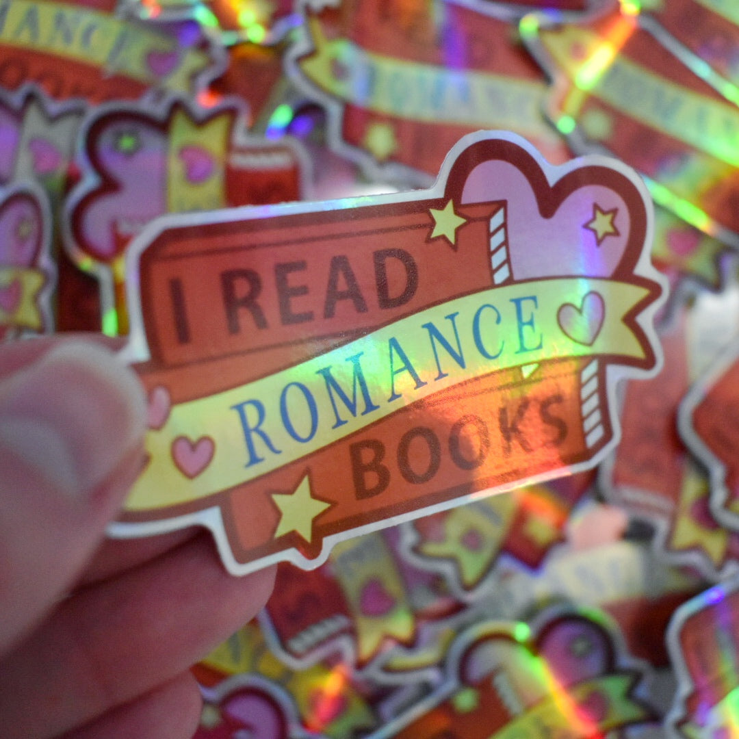 White hand holding sticker, tilted at an angle to show the holographic effect. Sticker design is a stack of red books with a big pink heart on right side of design. Small pink hearts and yellow stars are scattered on the design. A yellow banner across the middle of the design. The design reads I read romance books. Sticker has a white border all around the design. Background is blurry/distance of the same stickers scattered.