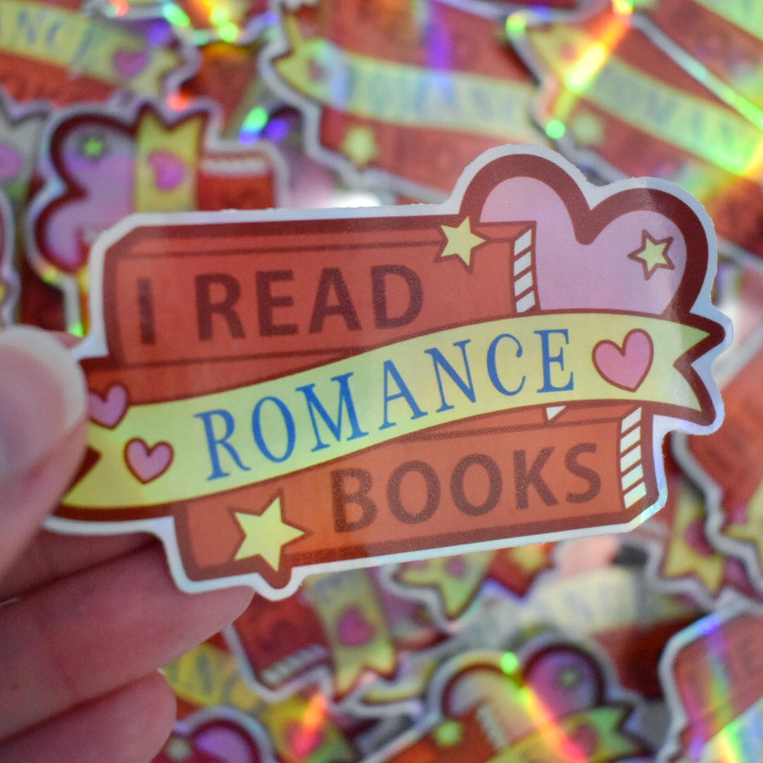White hand holding sticker. Sticker design is a stack of red books with a big pink heart on right side of design. Small pink hearts and yellow stars are scattered on the design. A yellow banner across the middle of the design. The design reads I read romance books. Sticker has a white border all around the design. Background is blurry/distance of the same stickers scattered.