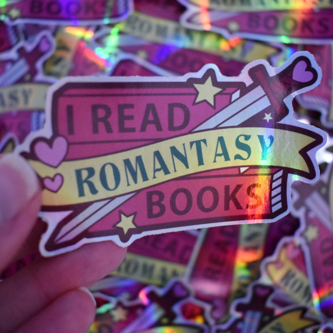 White hand holding sticker, tilted at an angle to show the holographic effect. Sticker design is a stack of dark pink books with a grey bladed sword, with a dark pink/reddish and pink hilt/handle. Small pink hearts and yellow stars are scattered on the design. A yellow banner across the middle of the design. The design reads I read romantasy books. Sticker has a white border all around the design. Background is blurry/distance of the same stickers scattered.