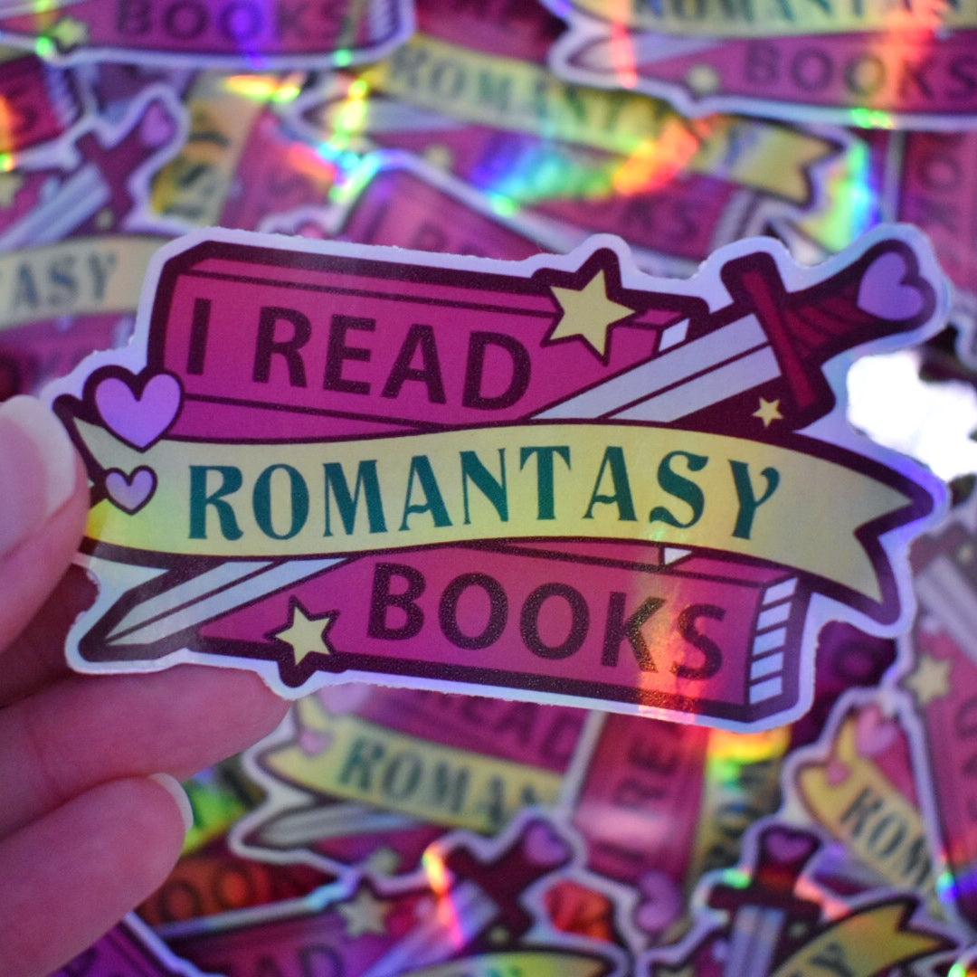 White hand holding sticker. Sticker design is a stack of dark pink books with a grey bladed sword, with a dark pink/reddish and pink hilt/handle. Small pink hearts and yellow stars are scattered on the design. A yellow banner across the middle of the design. The design reads I read romantasy books. Sticker has a white border all around the design. Background is blurry/distance of the same stickers scattered.