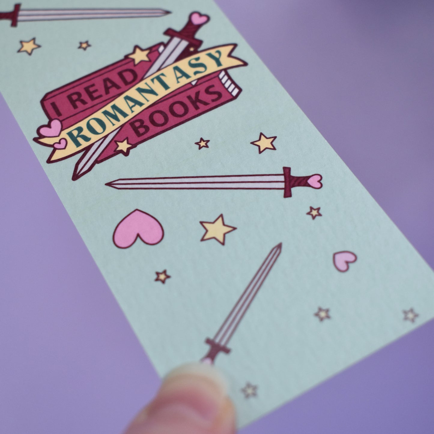 Close up of textured bookmark with 'I Read Romantasy Books' text on a small stack of books and banner, with a sword illustration, in the centre of the design. Smaller swords, hearts and stars fill the rest of the bookmark space. Held by a white hand against lilac background.