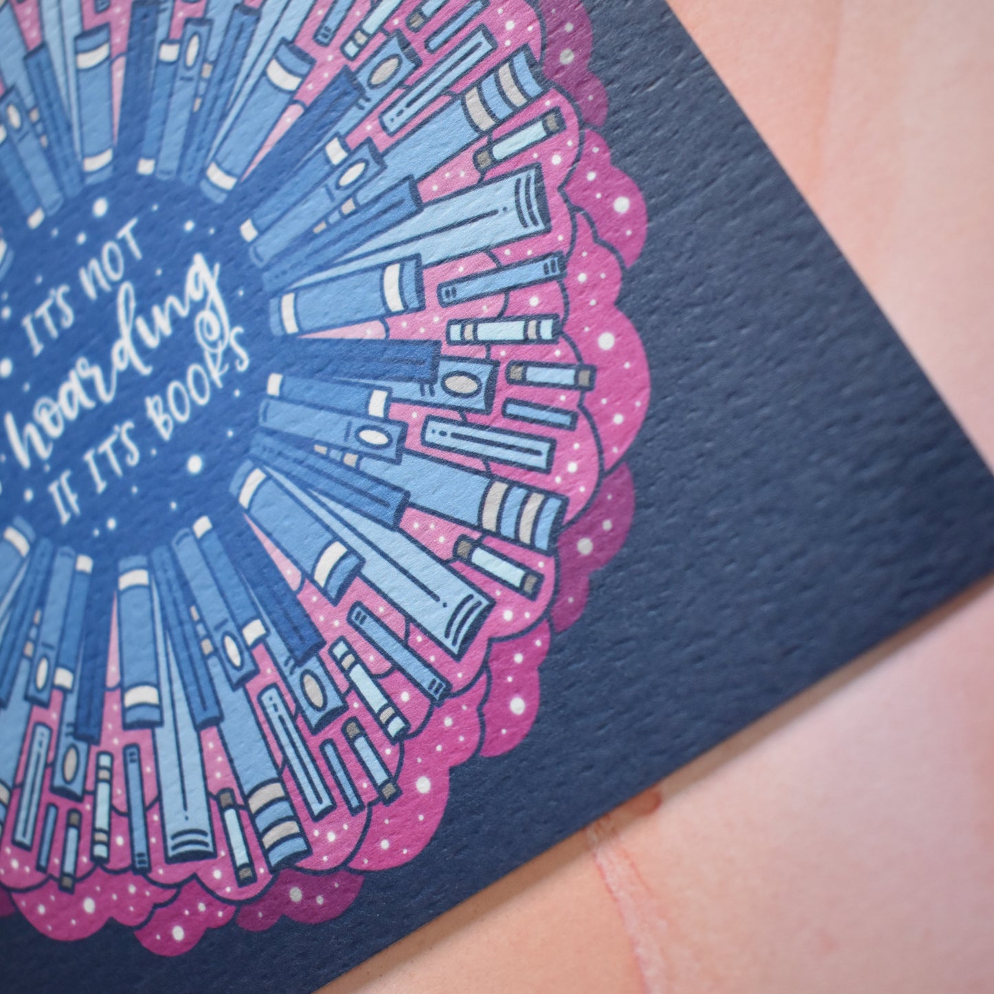 Close up of corner of design to show the texture of the print which is slightly raised. Square bookish print featuring a repeating pattern of blue bookspines and pink cloud/swirls with darker blue around the edge. Design reads 'it's not hoarding if it's books' in white/light blue at the centre of the design. Background is pink/lilac/peach watercolour style.
