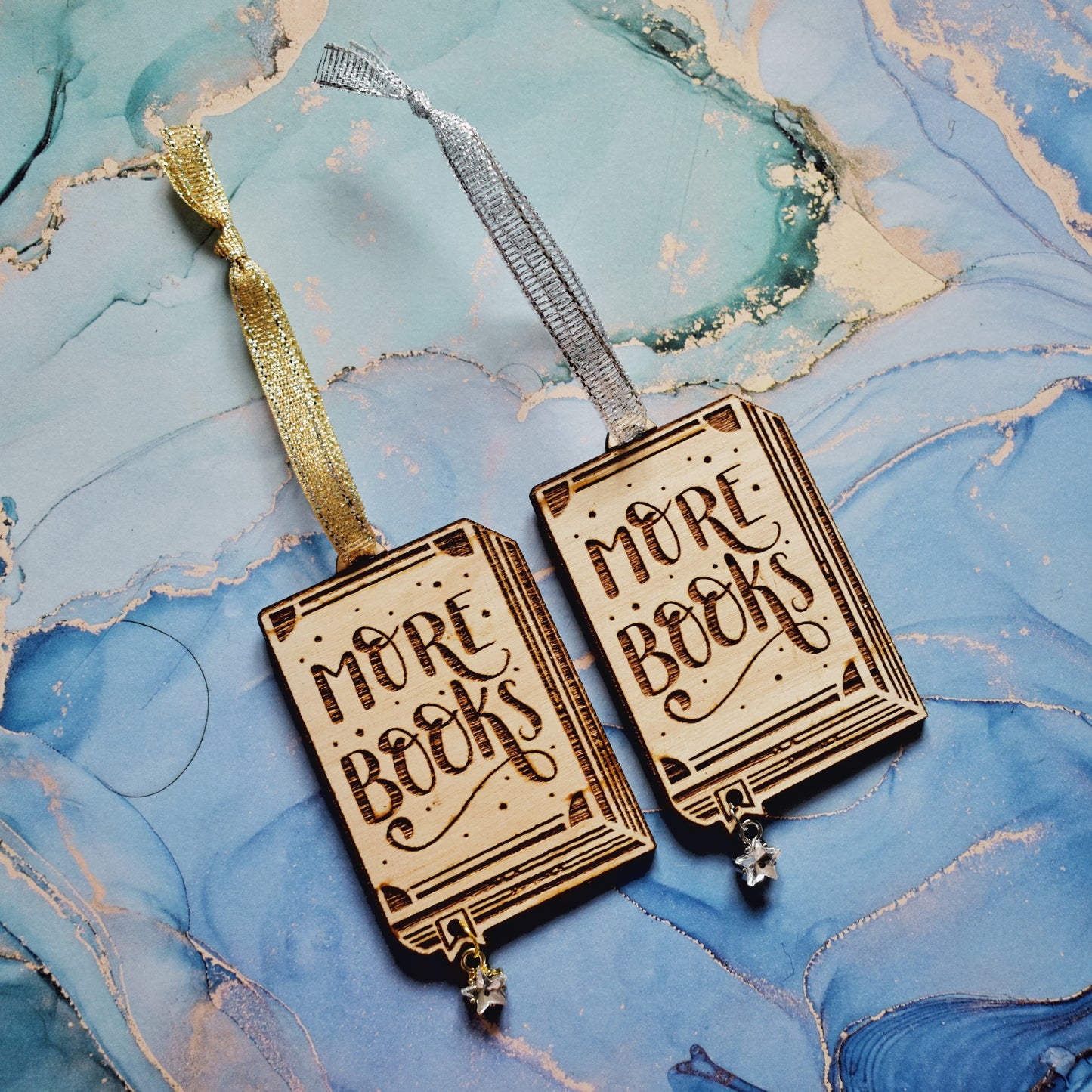 2 wooden ornaments next to each other, at an angle on a purple watercolour background. Ornaments are in the shape of a book with more books in handwritten style writing, showing the page edges at the side and bottom, with a small bookmark end sticking out. A hole in the bookmark end allows for the attachment of a small clear star gem. Left ornamant has a gold ribbon and gold loop on star gem. Right ornamant has a silver ribbon and silver loop on star gem.