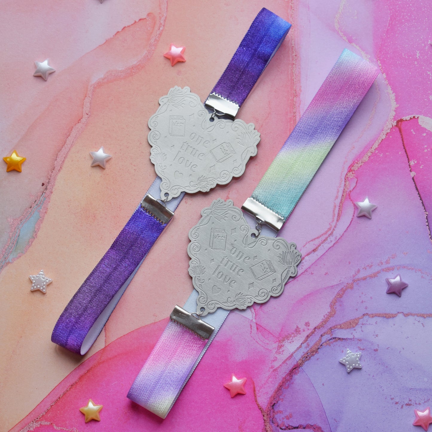 2 book bands at diagonal in the centre of image. Bottom bookband is pastel rainbow elastic with stainless steel engraved heart and book charm that reads one true love. Top bookband is purple galaxy elastic with stainless steel engraved heart and book charm that reads one true love. On a pale pink/peach/lilac watercolour background with different coloured bead stars scattered around.