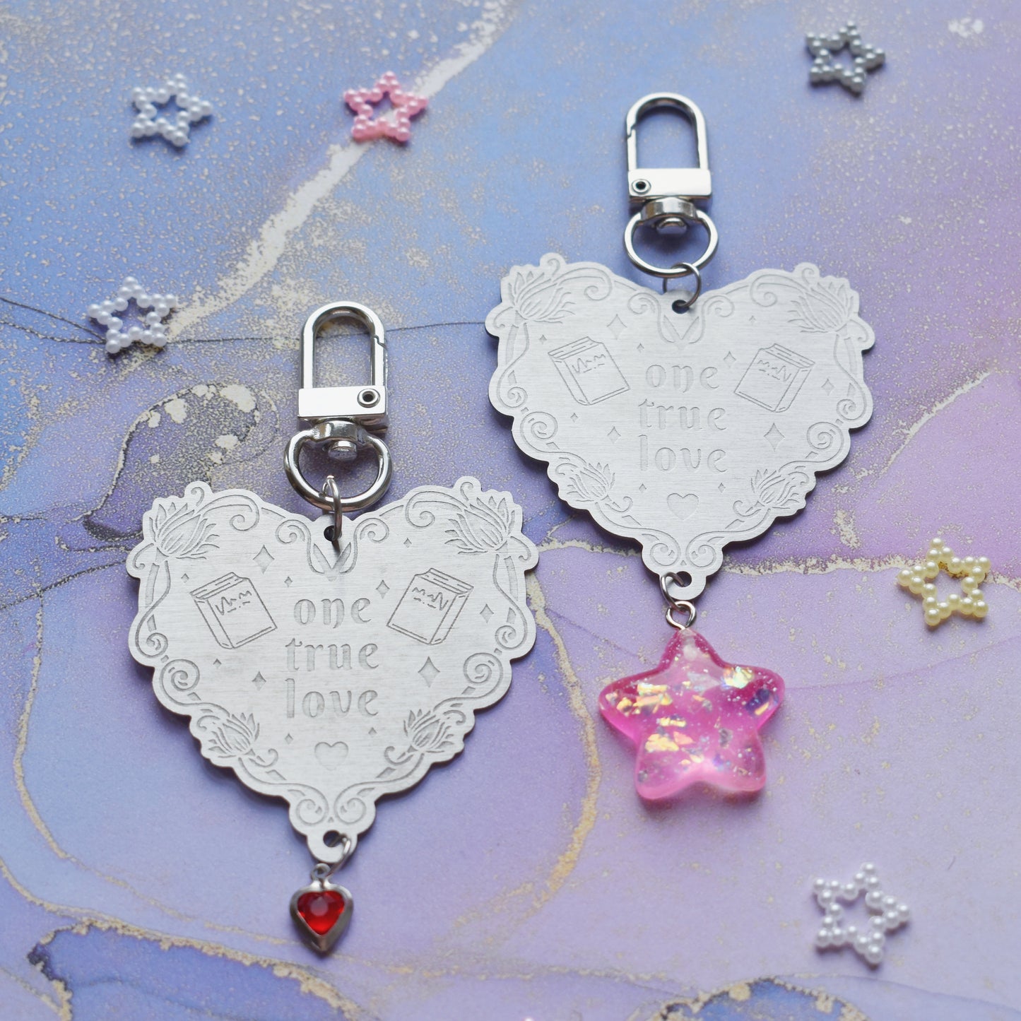 2 keyrings with stainless steel engraved heart and book charm that reads one true love in the centre of image. Left keyring has a small silver and gem red heart attached to charm. Right keyring has a pink glittery cabacon star attached to charm. On a pale blue/lilac watercolour background with different coloured bead stars scattered around.
