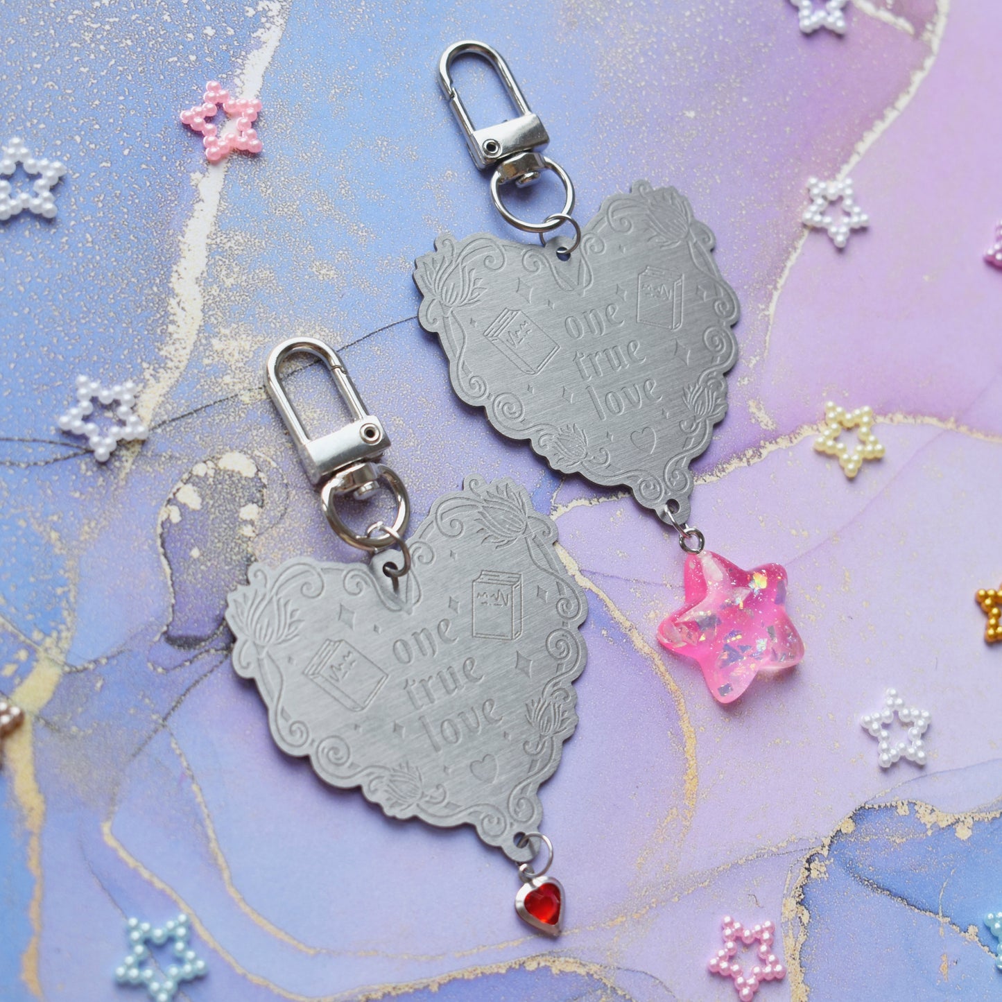 2 keyrings with stainless steel engraved heart and book charm that reads one true love at diagonal in the centre of image. Left keyring has a small silver and gem red heart attached to charm. Right keyring has a pink glittery cabacon star attached to charm. On a pale blue/lilac watercolour background with different coloured bead stars scattered around.