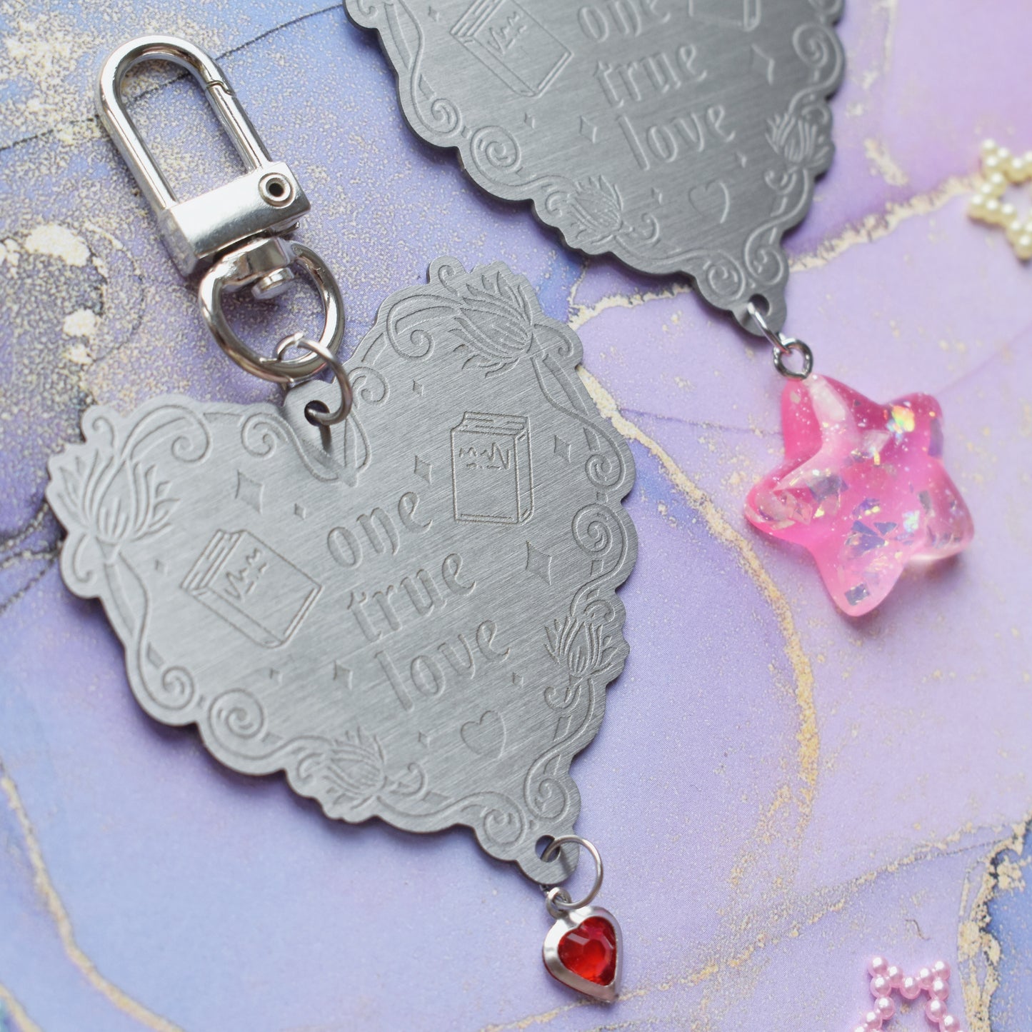 2 keyrings with stainless steel engraved heart and book charm that reads one true love at diagonal in the centre of image. Left keyring has a small silver and gem red heart attached to charm. Right keyring has a pink glittery cabacon star attached to charm. On a pale blue/lilac watercolour background with different coloured bead stars scattered around.