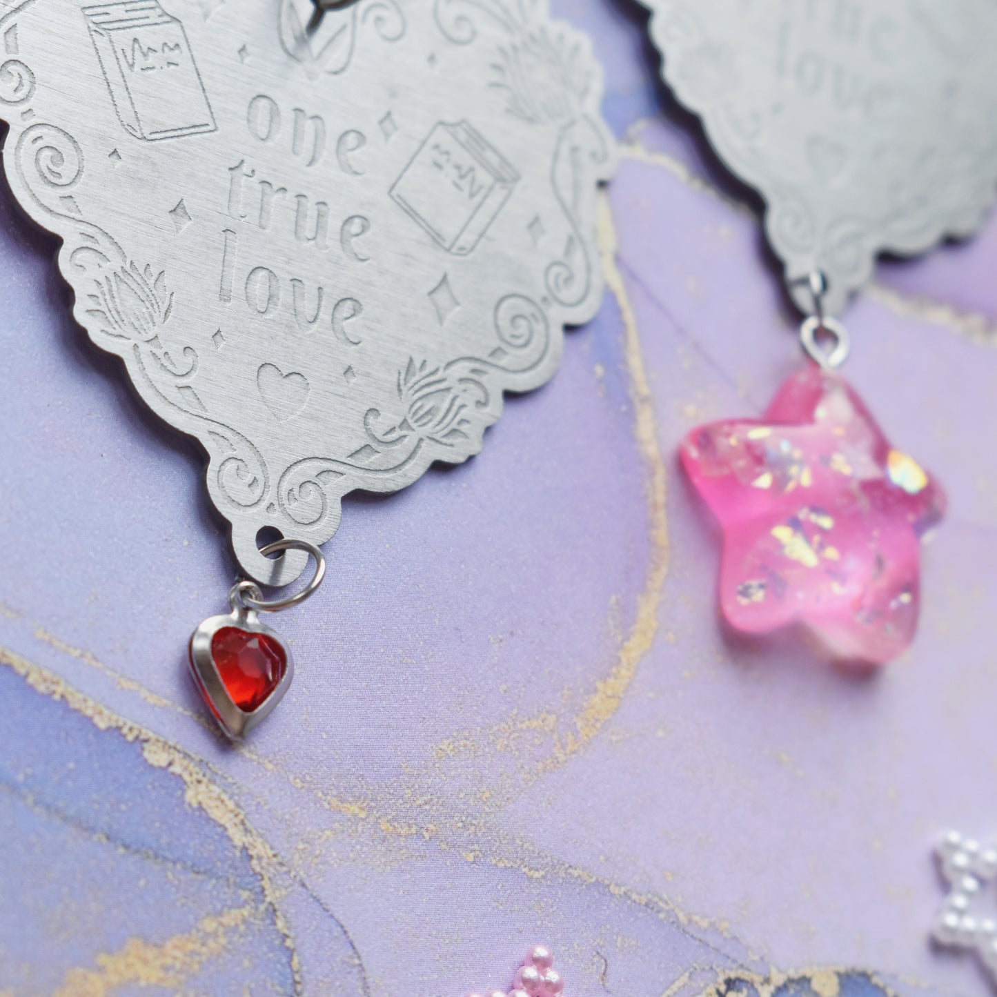 2 keyrings with stainless steel engraved heart and book charm that reads one true love at diagonal in the centre of image. Left keyring has a small silver and gem red heart attached to charm. Right keyring has a pink glittery cabacon star attached to charm. On a pale blue/lilac watercolour background with different coloured bead stars scattered around.