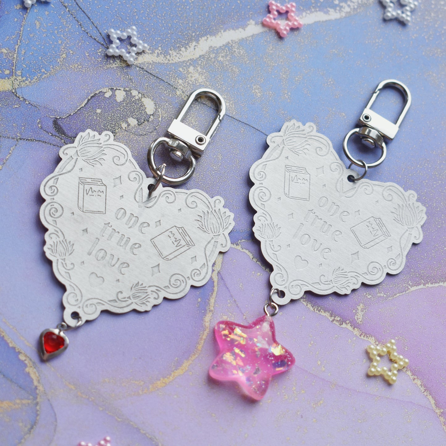 2 keyrings with stainless steel engraved heart and book charm that reads one true love at diagonal in the centre of image. Left keyring has a small silver and gem red heart attached to charm. Right keyring has a pink glittery cabacon star attached to charm. On a pale blue/lilac watercolour background with different coloured bead stars scattered around.