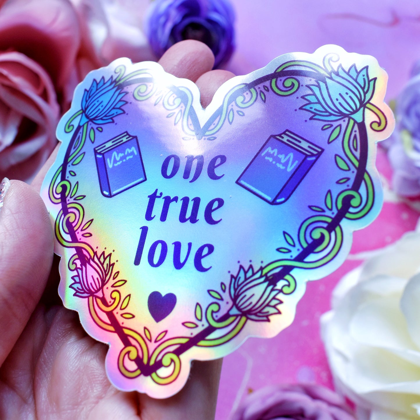 Purple heart-shaped bookish holographic sticker design features text 'one true love' with a floral and swirly outline with book covers either side of the text. Held at an angle by a white hand above a watercolour style background in blue, pink and purples with flowers around the edge of the photo.