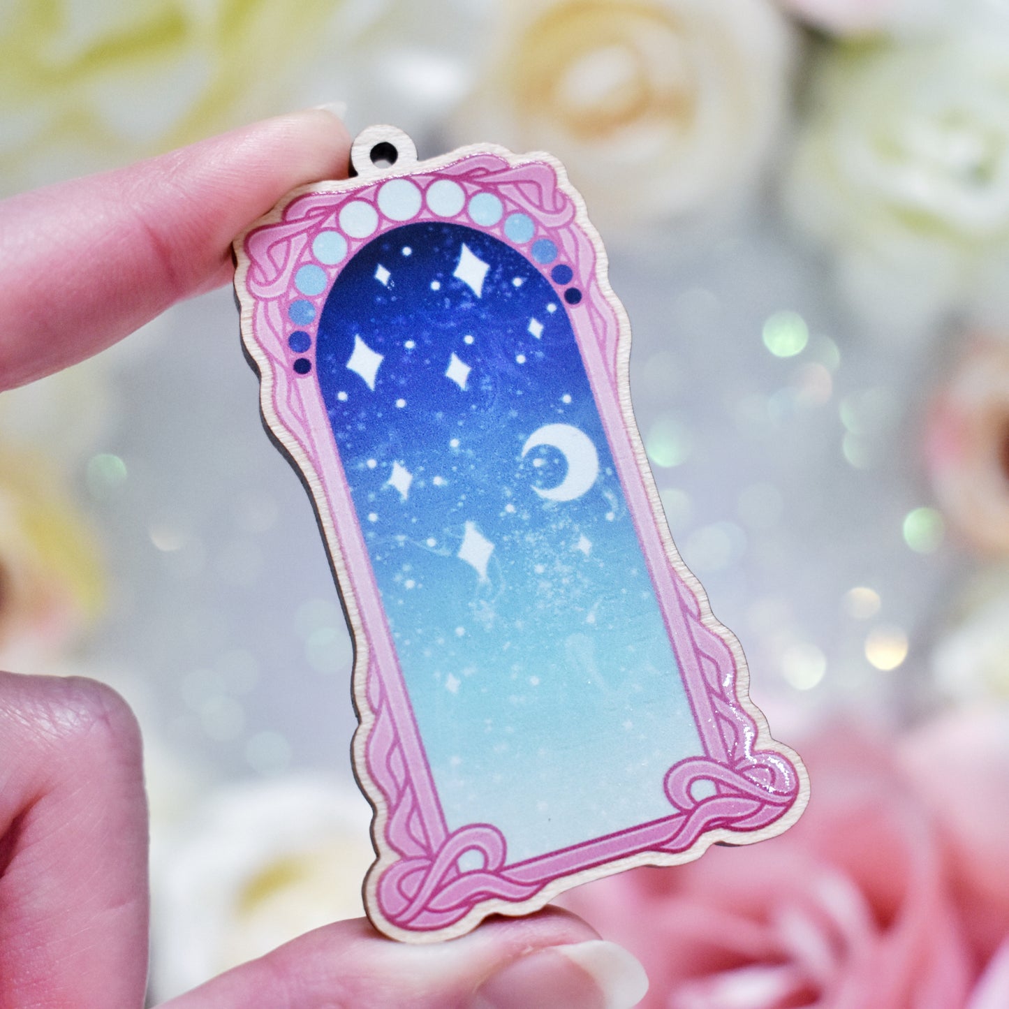 White hand holding wooden charm with top loop. Design has a pink detailed frame. Gradient colour pale teal through to blue with stars and a moon inside frame. Blurred fake flowers in white, yellow and pink are visible around edge of photo.
