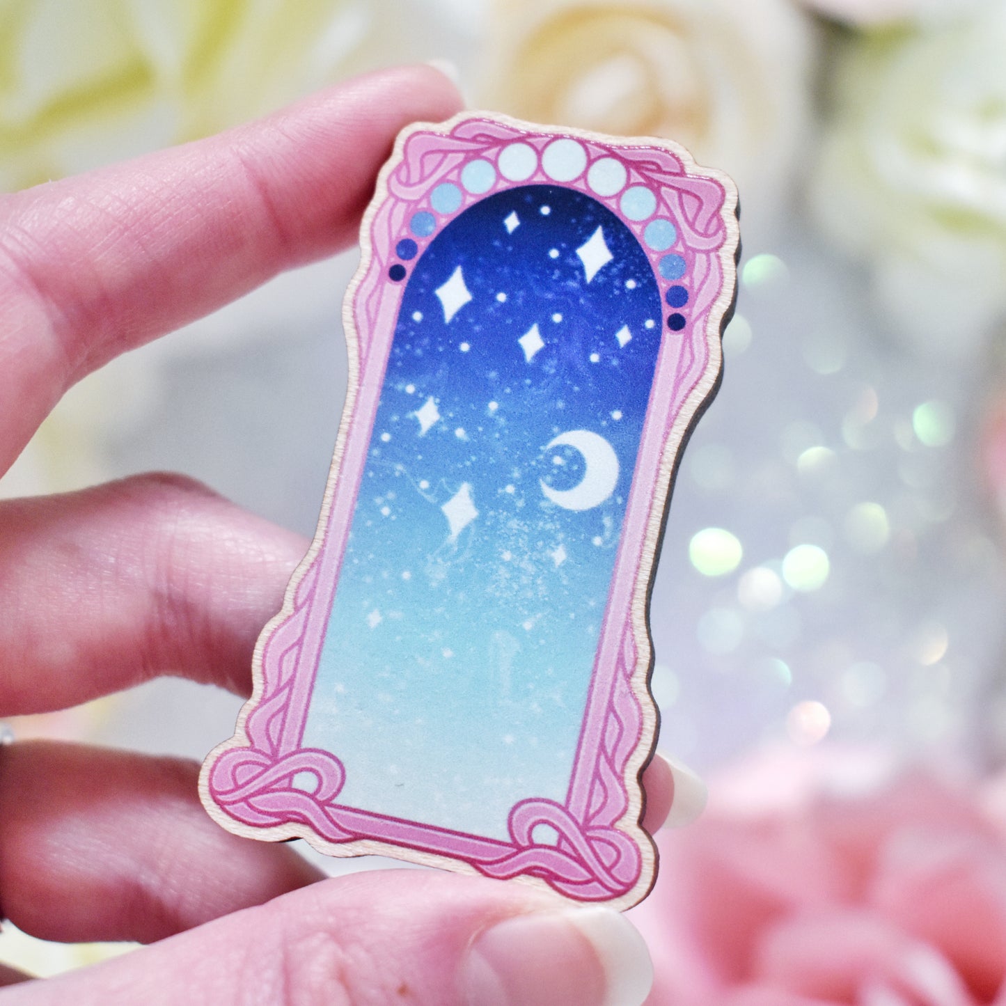 White hand holding wooden charm without top loop. Design has a pink detailed frame. Gradient colour pale teal through to blue with stars and a moon inside frame. Blurred fake flowers in white, yellow and pink are visible around edge of photo.