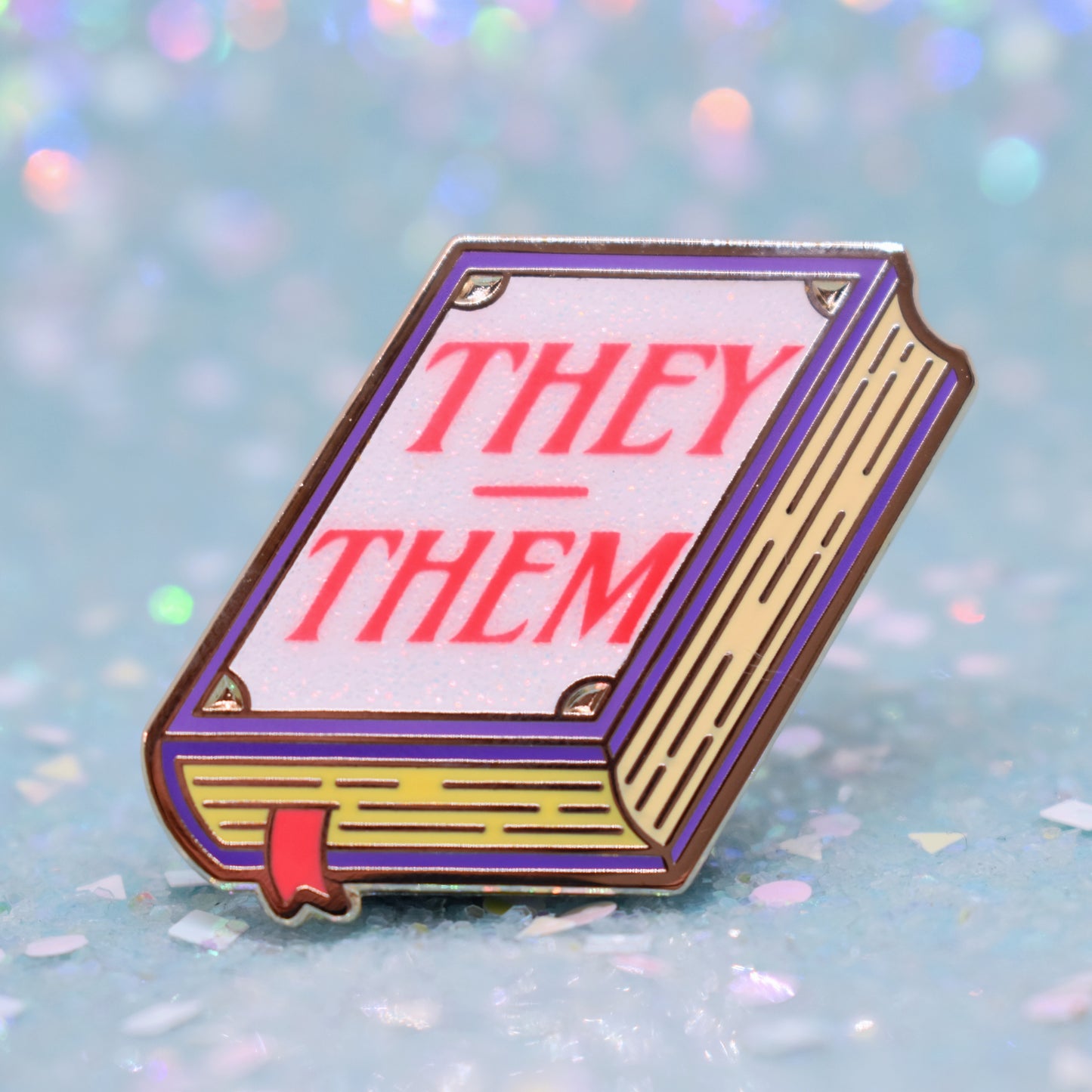 Book shaped enamel pin in gold metal, featuring purple, yellow and white glitter accents with 'THEY - THEM' in a bright red/pink. Background is a glittery icy blue.