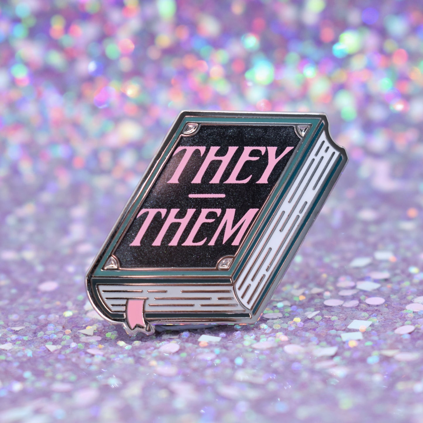 Book shaped enamel pin in silver metal, featuring teal, white and black shimmer accents with 'THEY - THEM' in a light pink. Background is a glittery lilac.