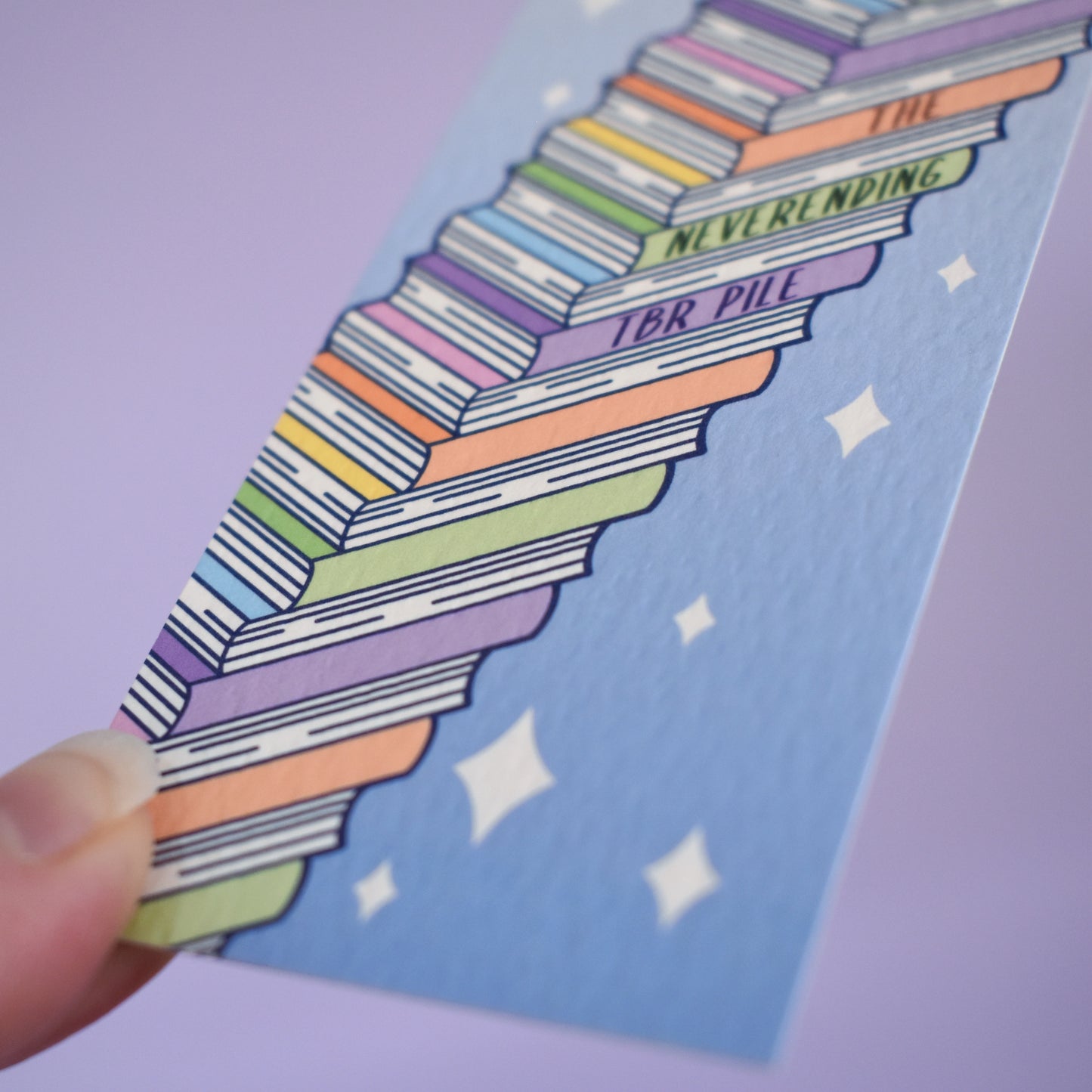 Close up of textured bookmark with 'The neverending TBR pile' on a angled stack of books running from bottom left to top right. Sparkles on a gradient blue background fill the rest of the bookmark space. Held by a white hand against lilac background.