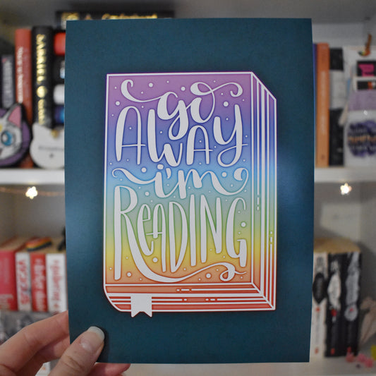 Teal illustration design with a rainbow book in the centre, facing forwards. 'Go away I'm reading' handwritten font in white ontop of the rainbow book. Held by a white hand in centre of image against a blurry bookcase with fairy lights.