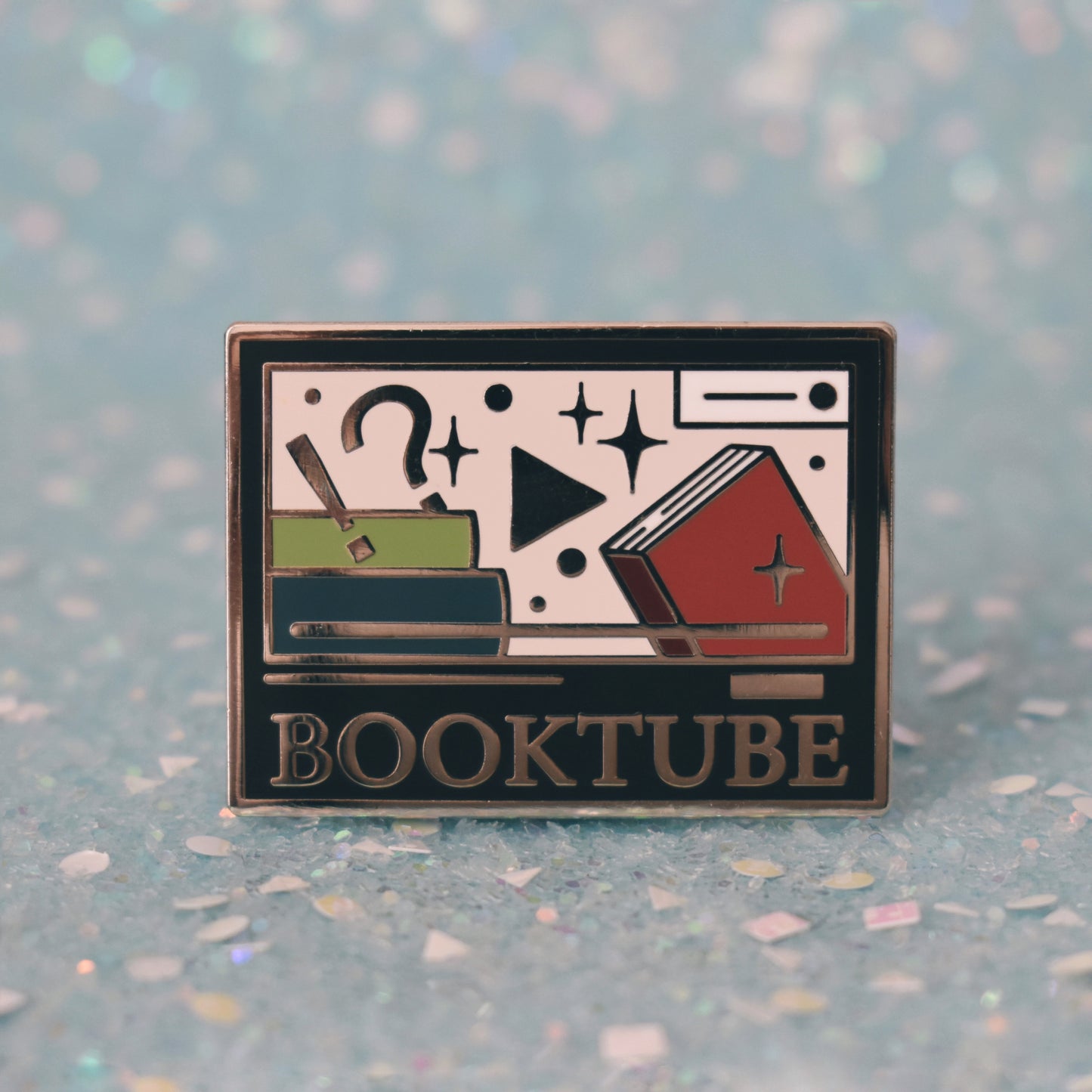 Silver metal enamel pin in centre of image. Enamel pin is rectangular and black, with a smaller cream rectangle inside which has multicoloured books, sparkles, a !? and play button. Under the smaller rectangle says booktube with a couple decorative lines. Background of photograph is sparkly teal.