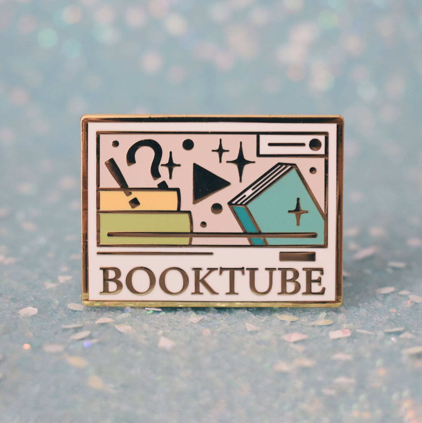 Gold metal enamel pin in centre of image. Enamel pin is rectangular and white, with a smaller cream rectangle inside which has multicoloured books, sparkles, a !? and play button. Under the smaller rectangle says booktube with a couple decorative lines. Background of photograph is sparkly teal.