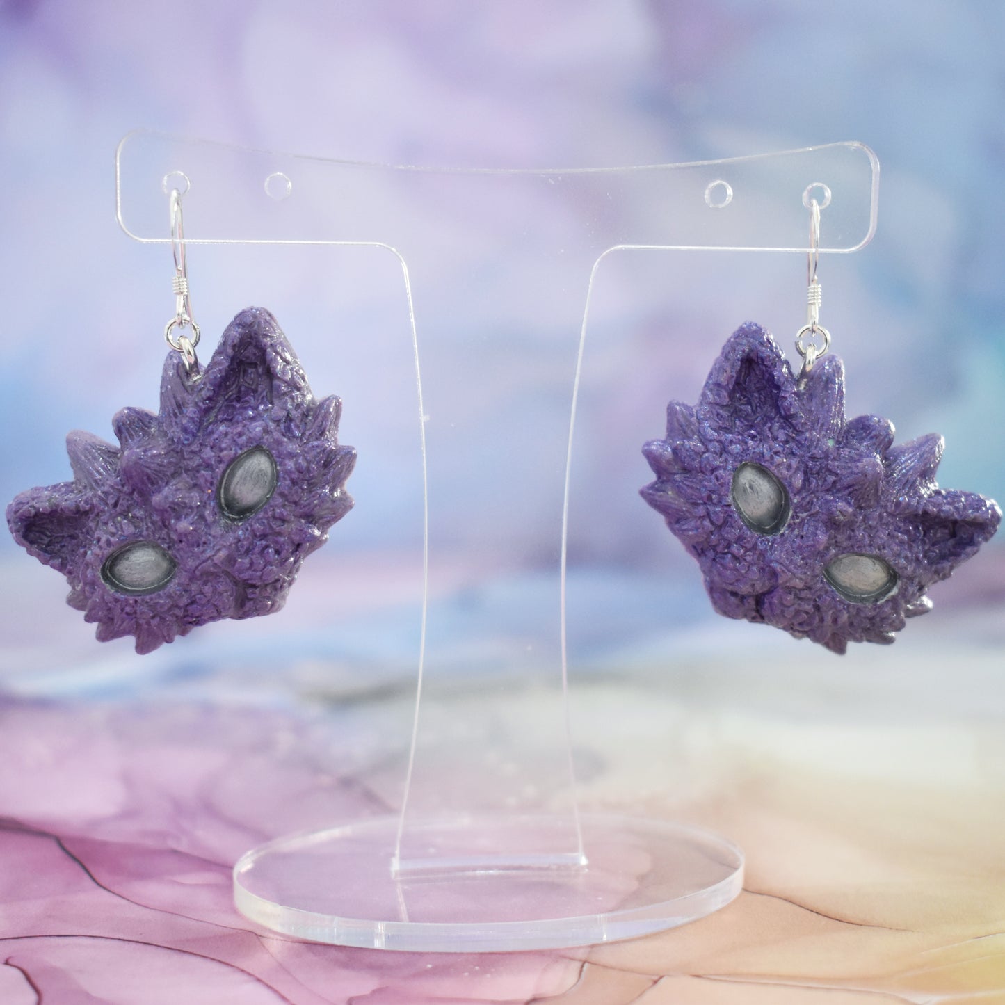 2 tilted purple dragon kitty heads with silver shimmer and silver eyes on silver earring hooks. Hanging from a clear mini earring display. Background is a soft multicolour watercolour style.