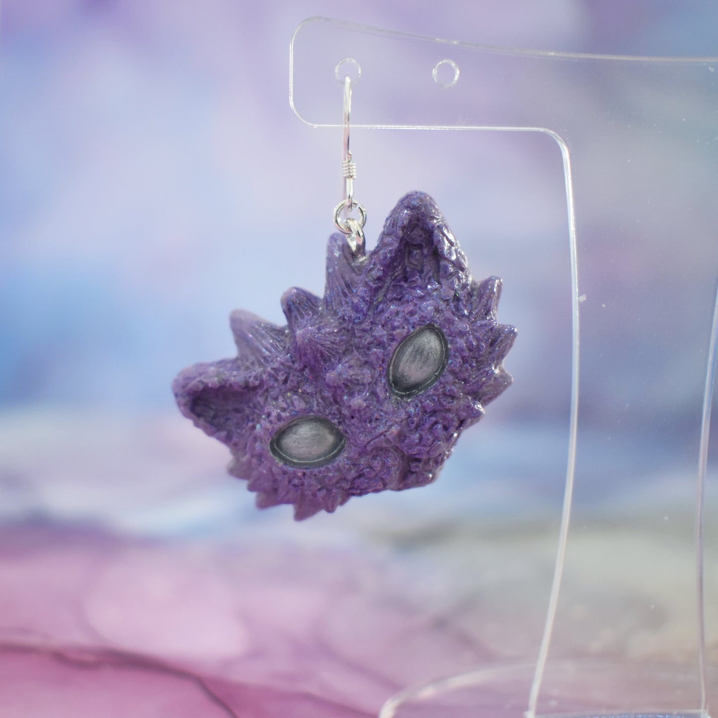 Left earring purple dragon kitty heads with silver shimmer and silver eyes on silver earring hooks. Hanging from a clear mini earring display. Background is a soft multicolour watercolour style.