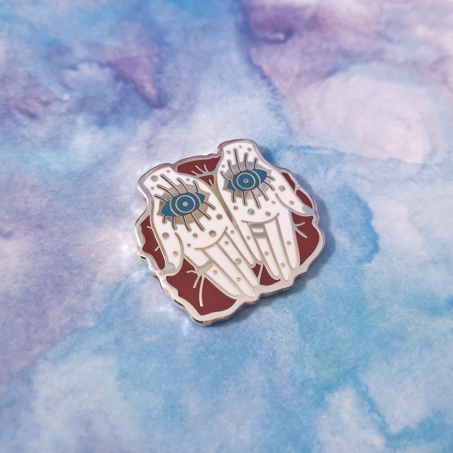 Photo shows an a silver metal enamel pin at the center of the image. A pair of palm up white hands with dot details and a blue open eye on each palm. The hands rest upon red petals. Background of photo is a blue/purple watercolour style.