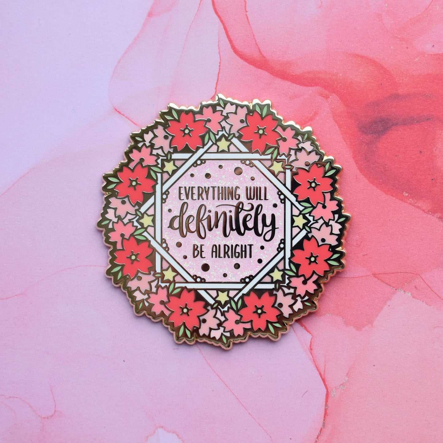 Gold metal enamel pin in centre of image. Enamel pin is roughly round with a ring of different sized stylized sakura flowers in different tones of pink around the rim. inside that is some white crossing over lines to make an 8 pointed star like shape with yellow stars and text everything will definitely be alright on a pink glitter centre. Background of photograph is pink smoke like style.