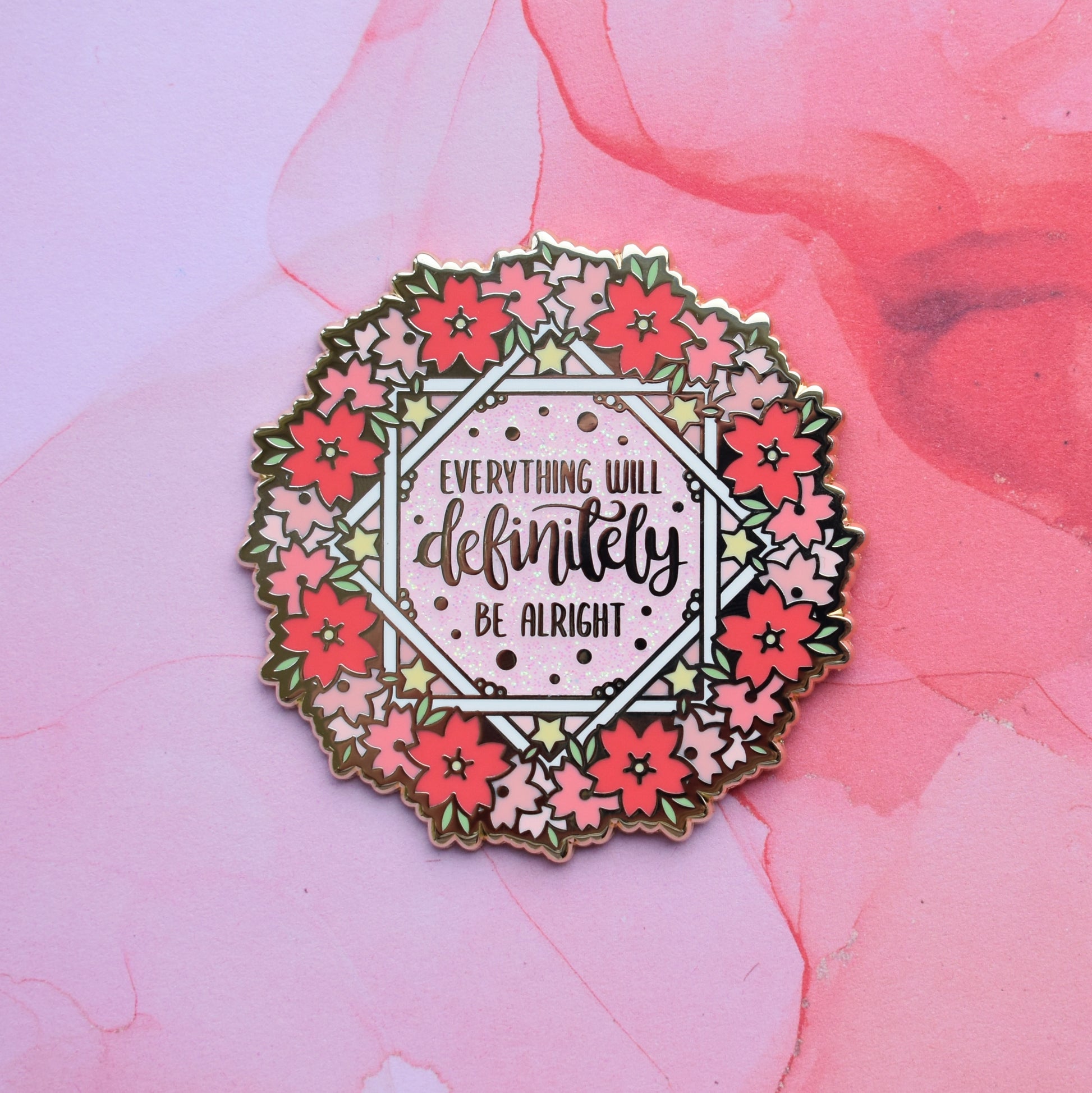 Gold metal enamel pin in centre of image. Enamel pin is roughly round with a ring of different sized stylized sakura flowers in different tones of pink around the rim. inside that is some white crossing over lines to make an 8 pointed star like shape with yellow stars and text everything will definitely be alright on a pink glitter centre. Background of photograph is pink smoke like style.