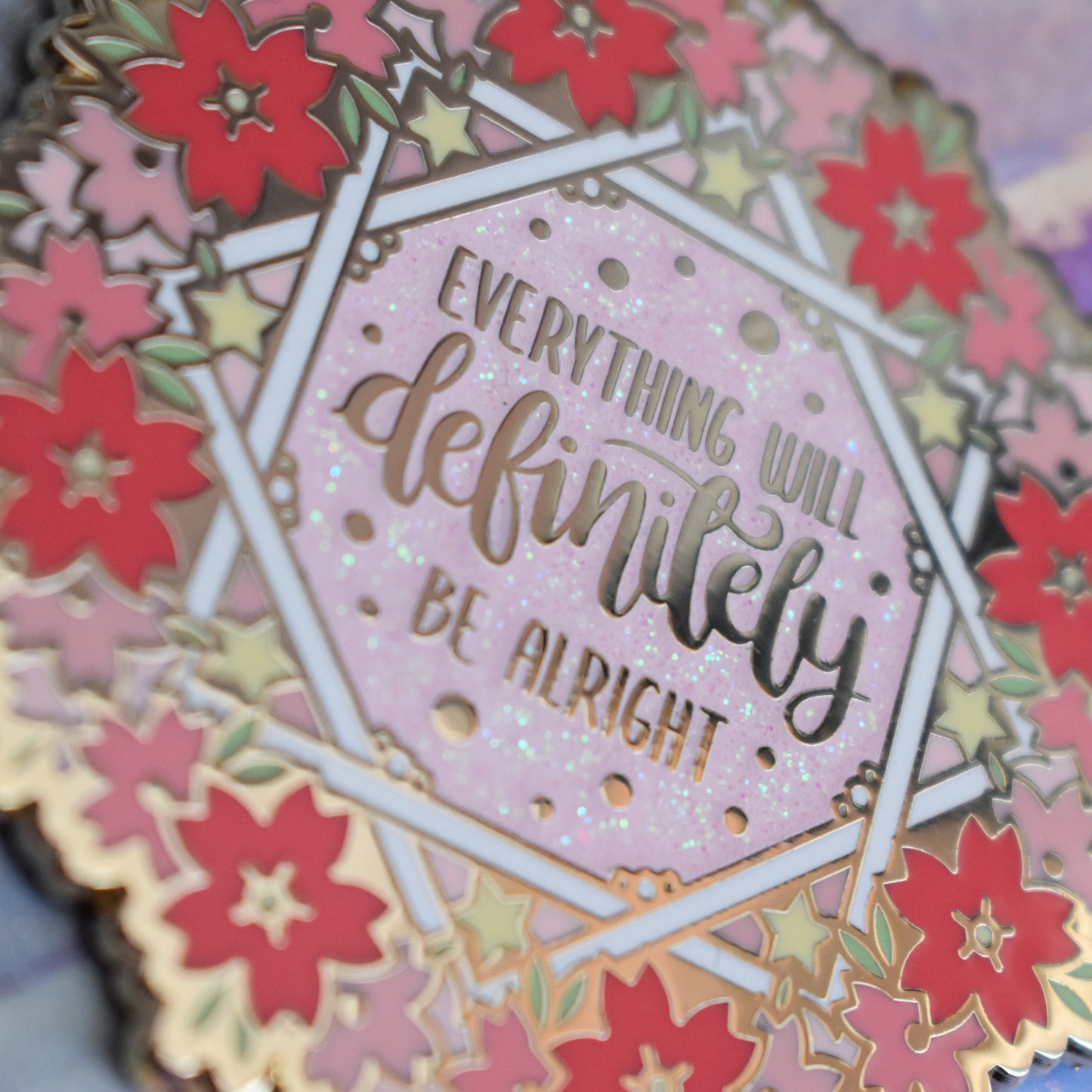 Close up of gold metal enamel pin in centre of image. Enamel pin is roughly round with a ring of different sized stylized sakura flowers in different tones of pink around the rim. inside that is some white crossing over lines to make an 8 pointed star like shape with yellow stars and text everything will definitely be alright on a pink glitter centre.
