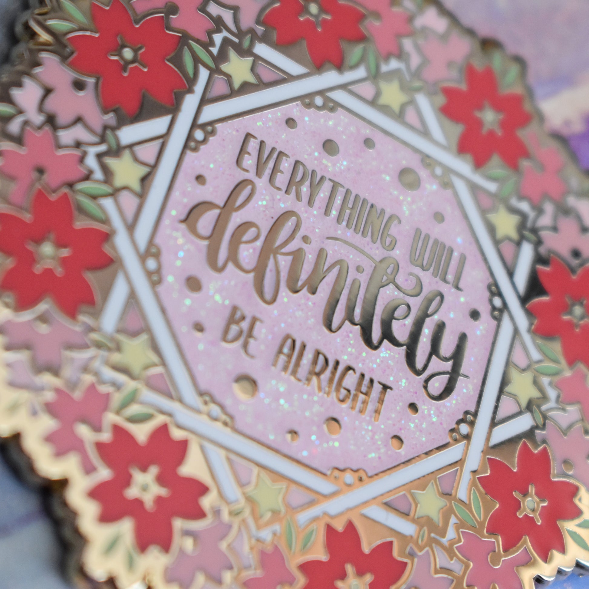 Close up of gold metal enamel pin in centre of image. Enamel pin is roughly round with a ring of different sized stylized sakura flowers in different tones of pink around the rim. inside that is some white crossing over lines to make an 8 pointed star like shape with yellow stars and text everything will definitely be alright on a pink glitter centre.