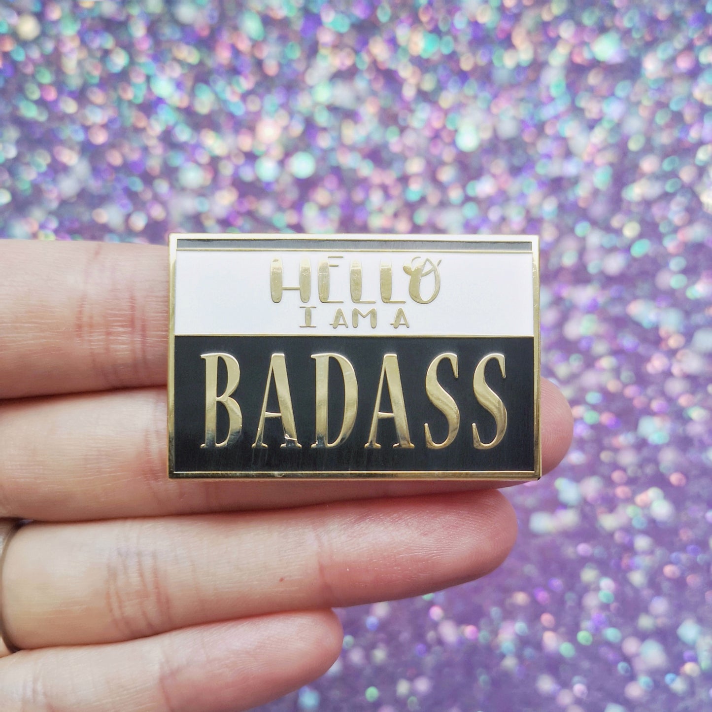 White hand holding rectangle gold metal enamel pin. Black background with a white band across most of top half. Hello I am a badass written across design. Badass in a large serif font. Background of photo is a blurry sparkly purple.