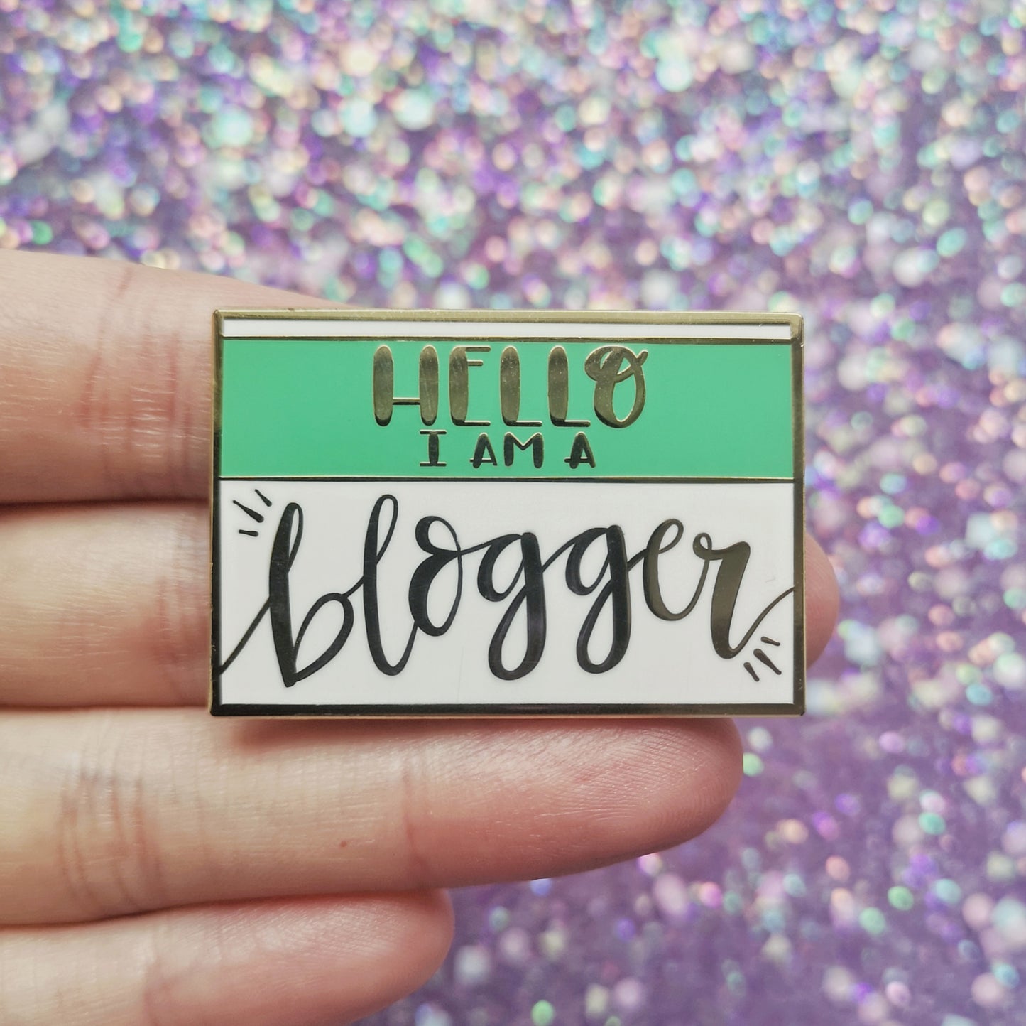 White hand holding rectangle gold metal enamel pin. White background with a green band across most of top half. Hello I am a blogger written across design. Blogger in a large loopy handwritten style font. Background of photo is a blurry sparkly purple.