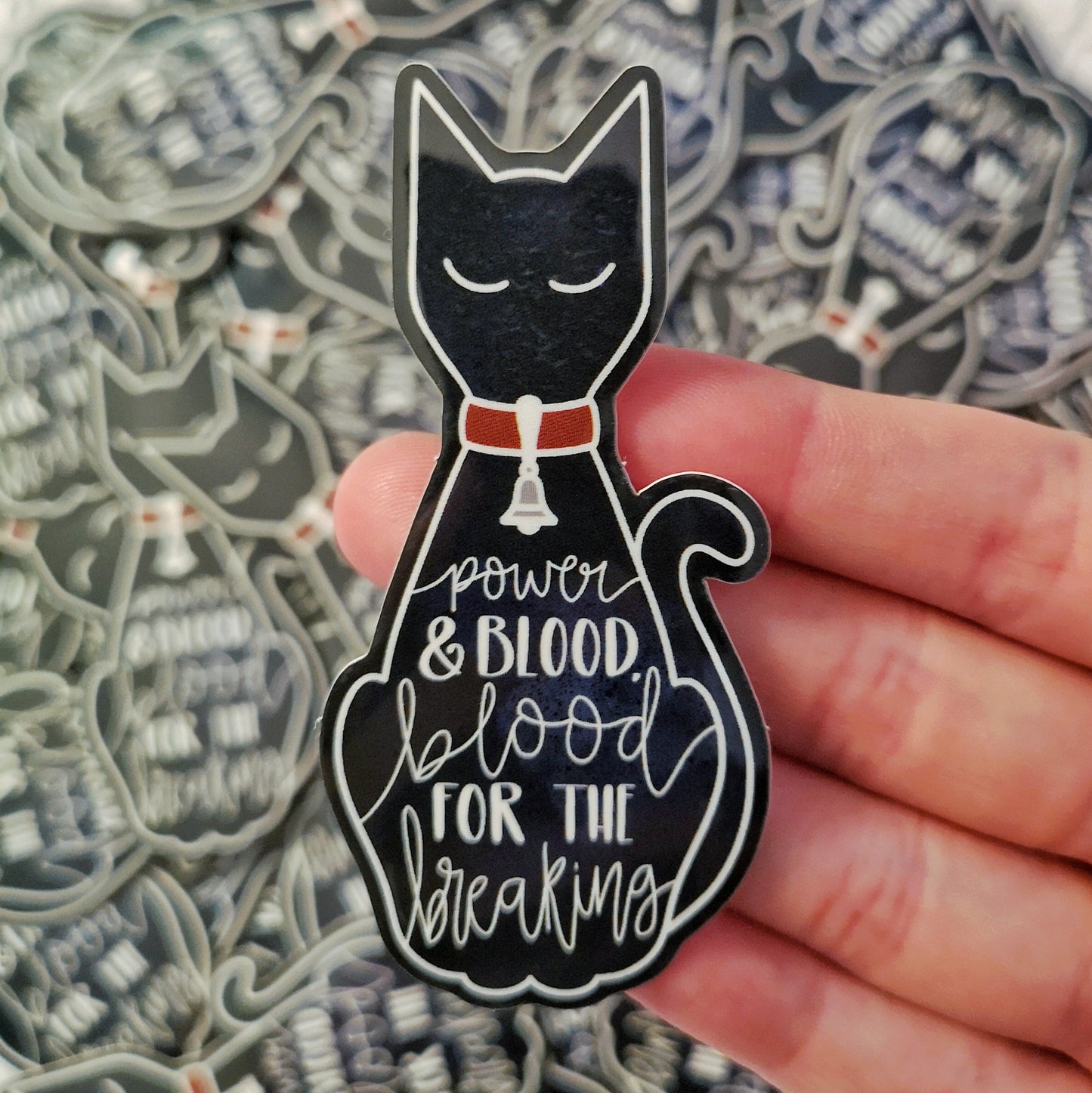 White hand holding sticker. Black feline shape, no visable paws or eye/ear details. A red collar with tiny bell around it's neck. The words power & blook, blood for the breaking across the design in white. Background is blurry/distance of same stickers.