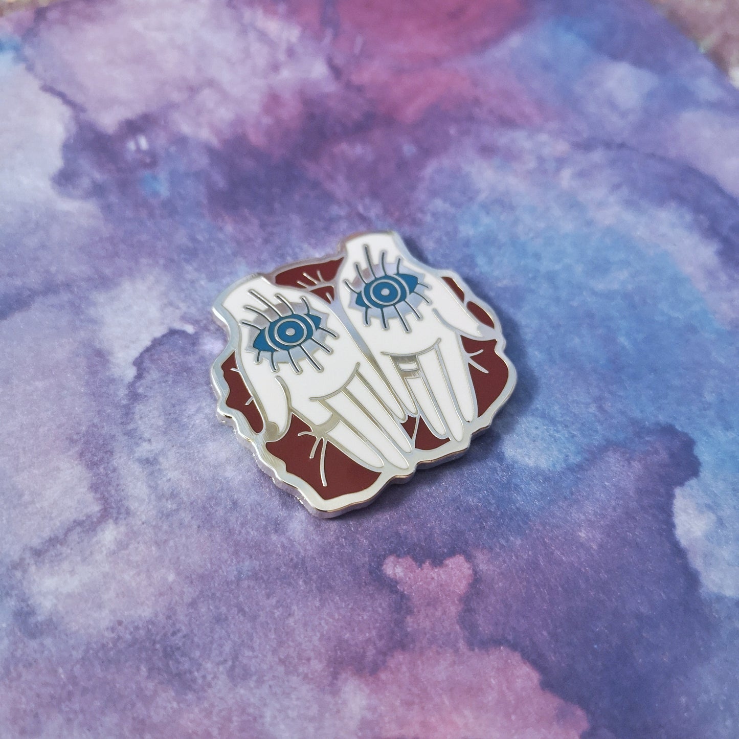Photo shows an a silver metal enamel pin at the center of the image. A pair of palm up white hands with a blue open eye on each palm. The hands rest upon red petals. Background of photo is a blue/purple watercolour style.