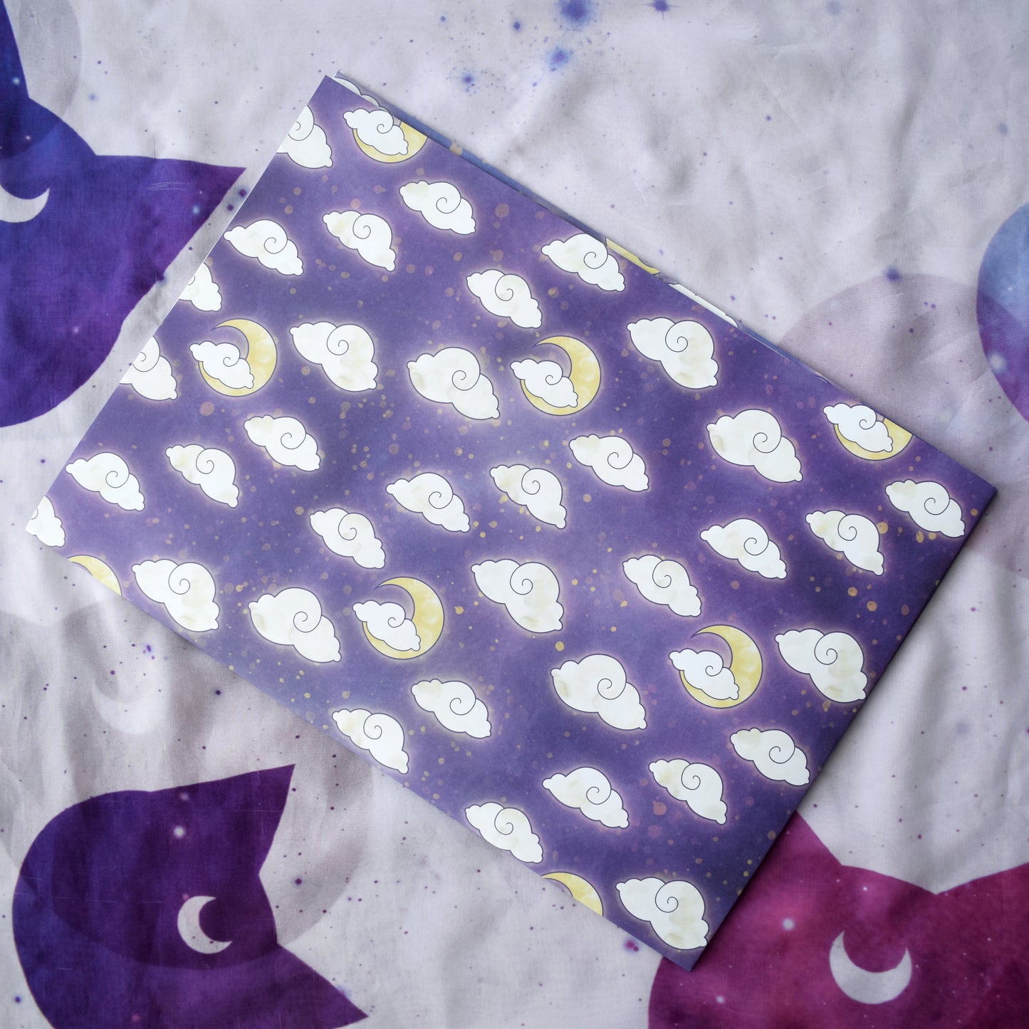 Folded down wrapping paper into a4 size, tilted in the middle of image. Wrapping paper is purple with lighter wisps and yellowish splatters. Features a repeating apptern of big fluffy white/cream clouds and yellow moons. Background is a off white/lilac with purple cat faces and purple sparkles.