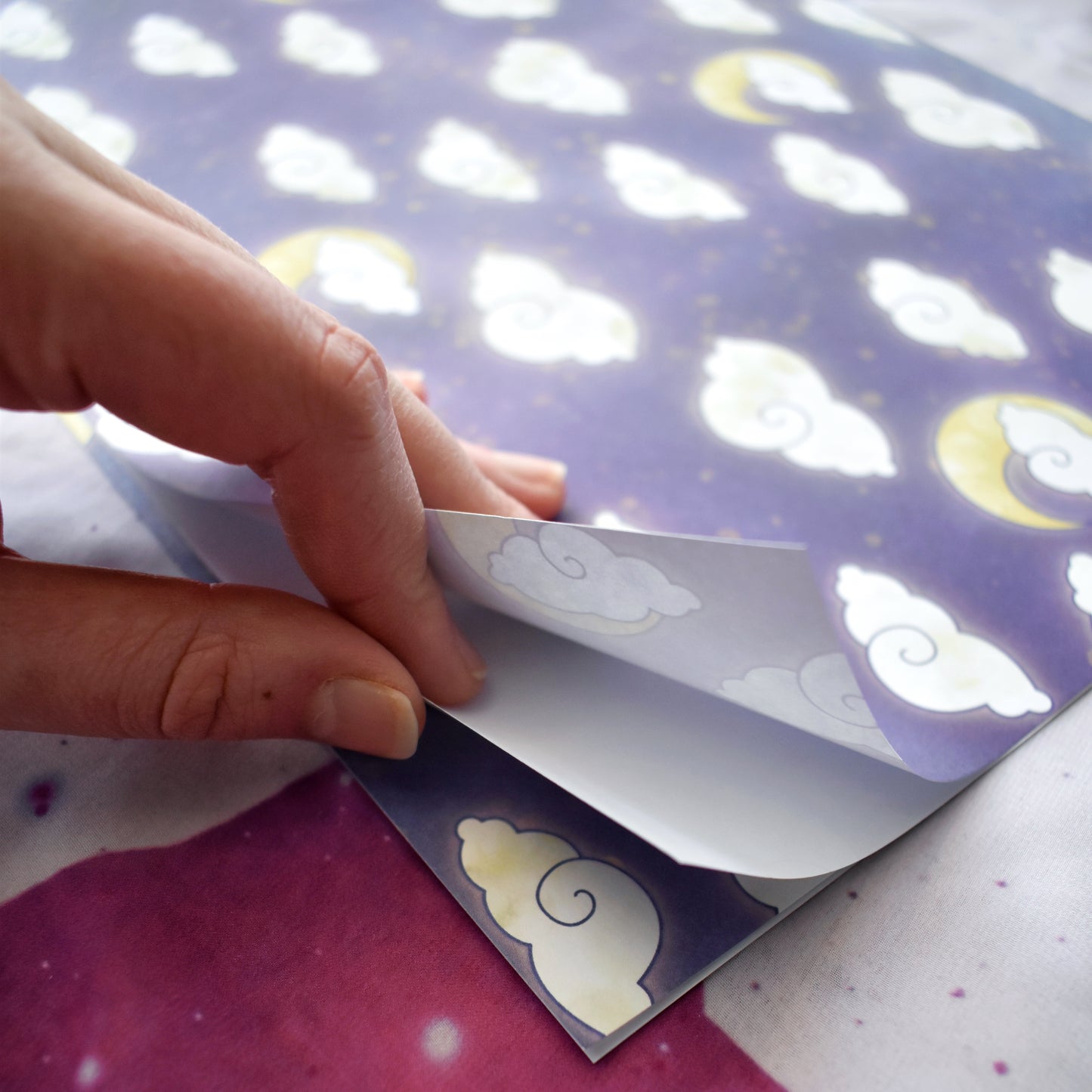 Close up of white hand holding open the layers of the folded down wrapping paper (into a4 size) Wrapping paper is purple with lighter wisps and yellowish splatters. Features a repeating apptern of big fluffy white/cream clouds and yellow moons. Background is a off white/lilac with purple cat faces and purple sparkles.