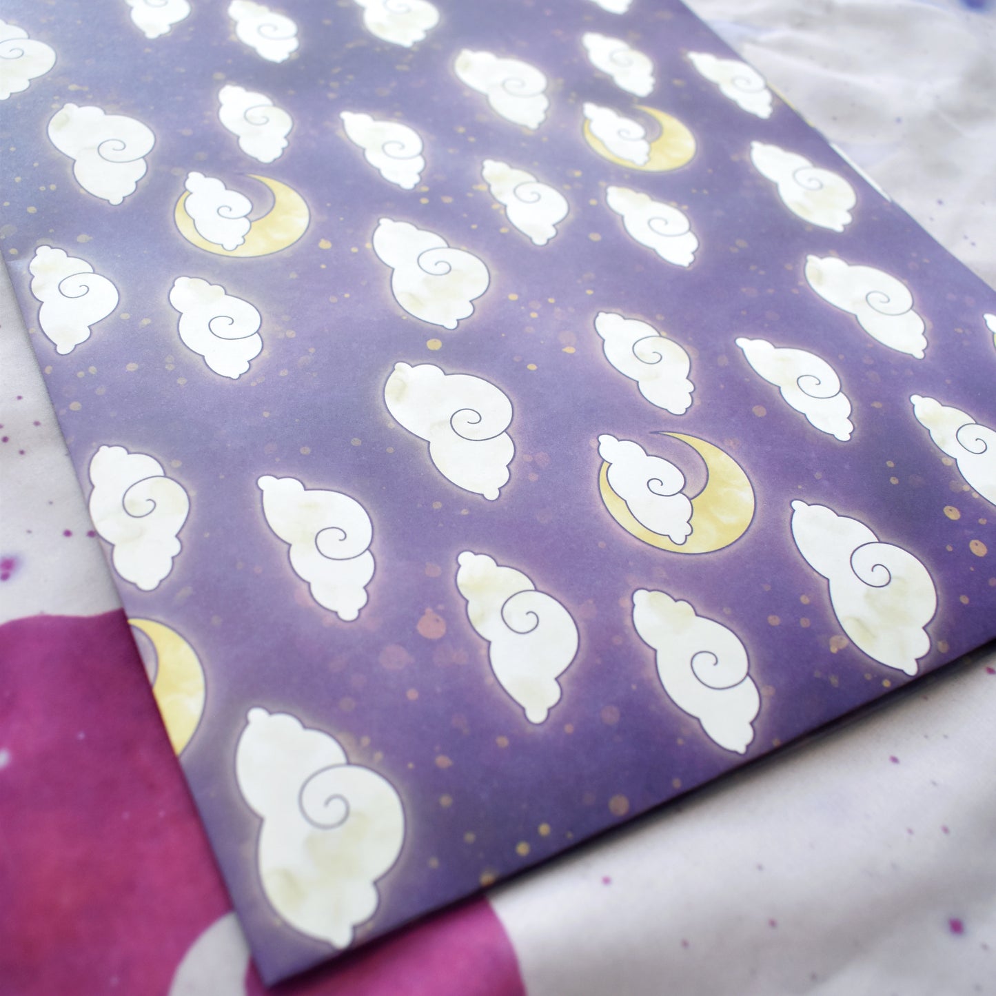 Close up of olded down wrapping paper into a4 size. Wrapping paper is purple with lighter wisps and yellowish splatters. Features a repeating apptern of big fluffy white/cream clouds and yellow moons. Background is a off white/lilac with purple cat faces and purple sparkles.