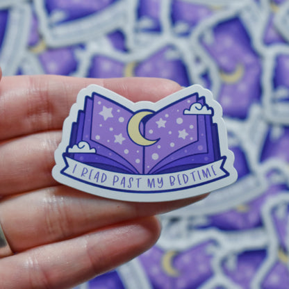 White hand holding sticker. Sticker design is an open book with a purple gradient, with a yellow moon top middle and white dots/stars/clouds spread across the design. A white banner at the bottom reads I read past my bedtime. Sticker has a white border. Background is blurry/distance of same stickers.